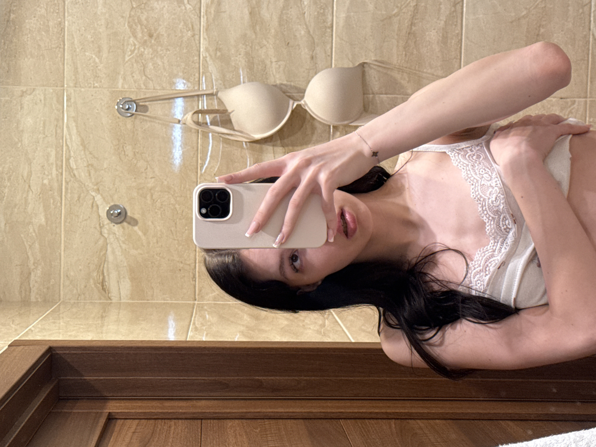 In a bathroom with beige tiled walls and a wooden door, a young woman with long dark hair takes a selfie in a mirror. She is wearing a white camisole with lace trim. Her right hand holds a white smartphone to capture the image, while her left hand is placed on her chest. A beige bra hangs on hooks on the wall behind her. The woman is looking directly into the camera with a neutral expression. The scene is indoors and appears to be in a private residence.