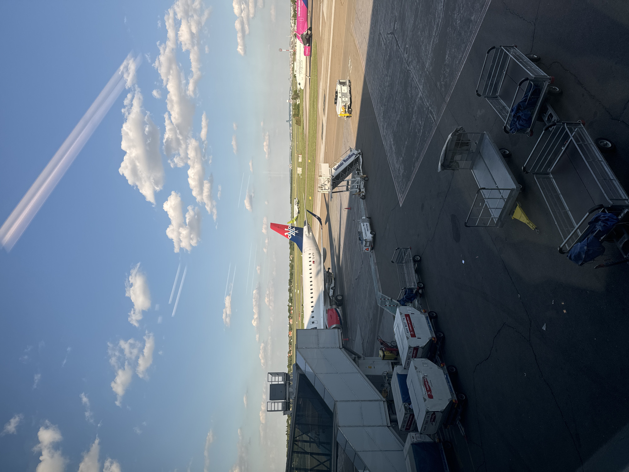 This image shows an airport scene, likely taken from an elevated position inside a terminal. The location is most likely the city of Surcin, Urban Municipality, Serbia given the airplanes and ground vehicles in the photo. The scene includes an aircraft, a vehicle with the name "Goldair Handling", luggage carts, and several ground service vehicles with "TURKISH AIR" written on their side. The sky is visible with white clouds.
