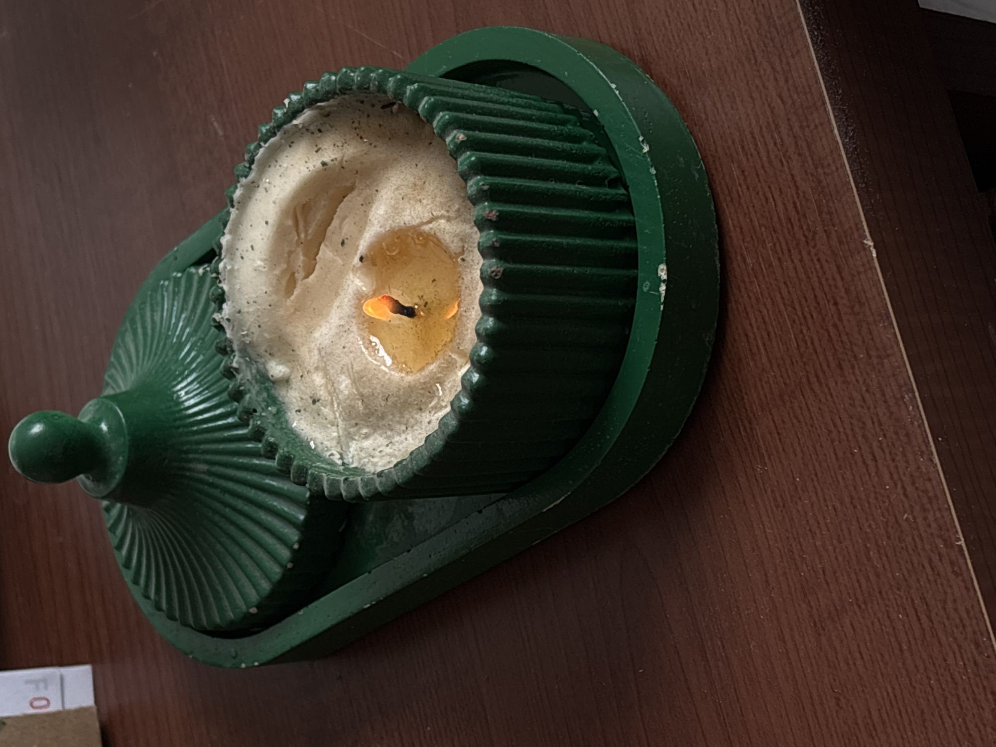 A green candle holder containing a burning candle sits on a dark brown, wood-grain surface. The candle holder has a circular base with a raised, ribbed edge, and a smaller, ribbed cover with a knob on top. The candle itself is mostly burnt down, with melted wax visible around the wick and flame. The scene is not identifiable to Santa Clara Ocoyucan, Mexico.