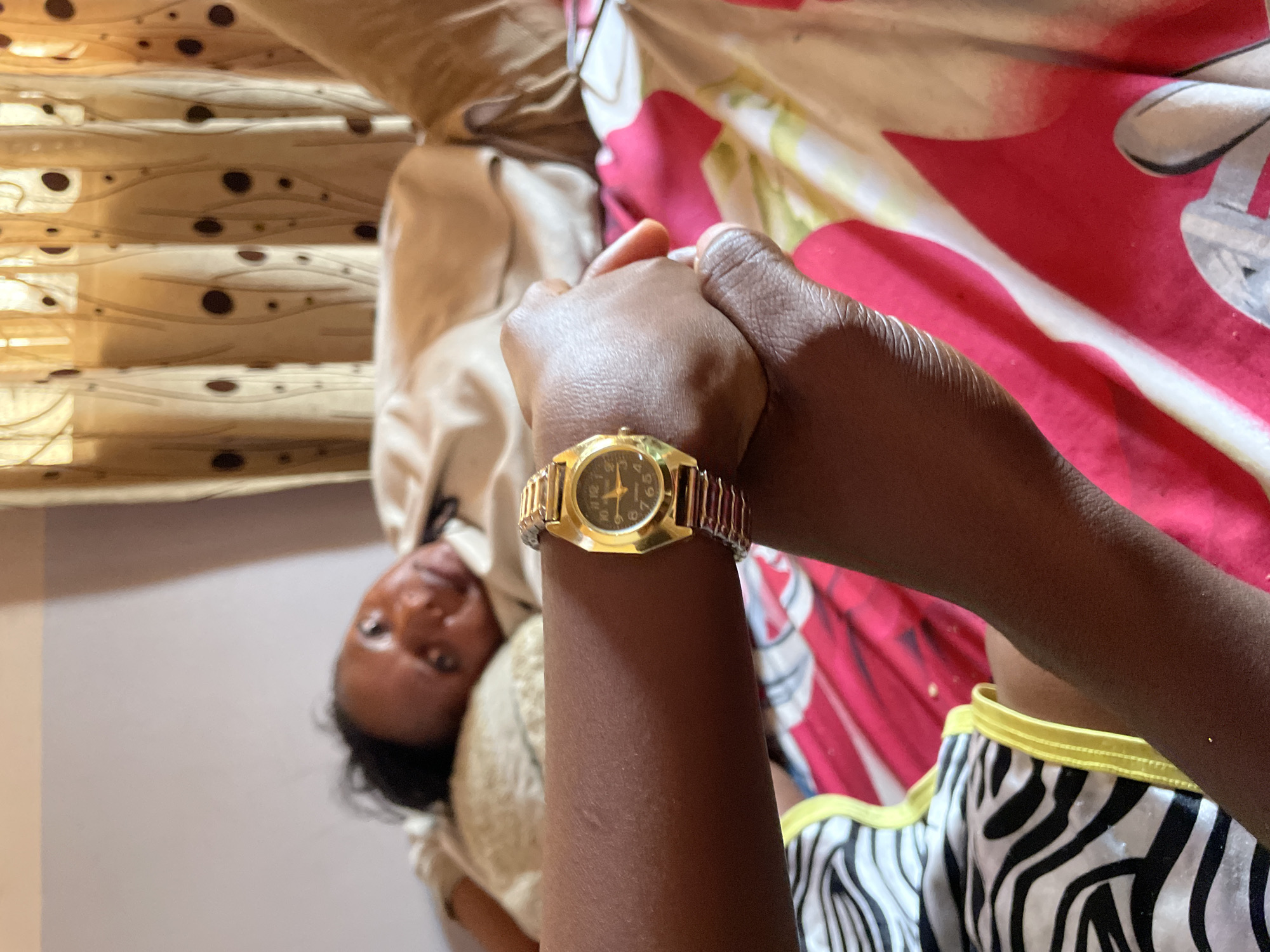 A photograph taken indoors shows a person lying down, seen from an upside-down perspective, with a gold-colored wristwatch prominently displayed on their wrist. The person is wearing clothing with a zebra-striped pattern and yellow trim, suggesting pajama bottoms or shorts. The hands of the person are clasped. The background includes bedding with a mix of patterns and colors, including a light tan or beige fabric with a pattern of spots and another fabric with red and white designs. The location of Jos, Nigeria, is not discernible from the visual elements within the image.