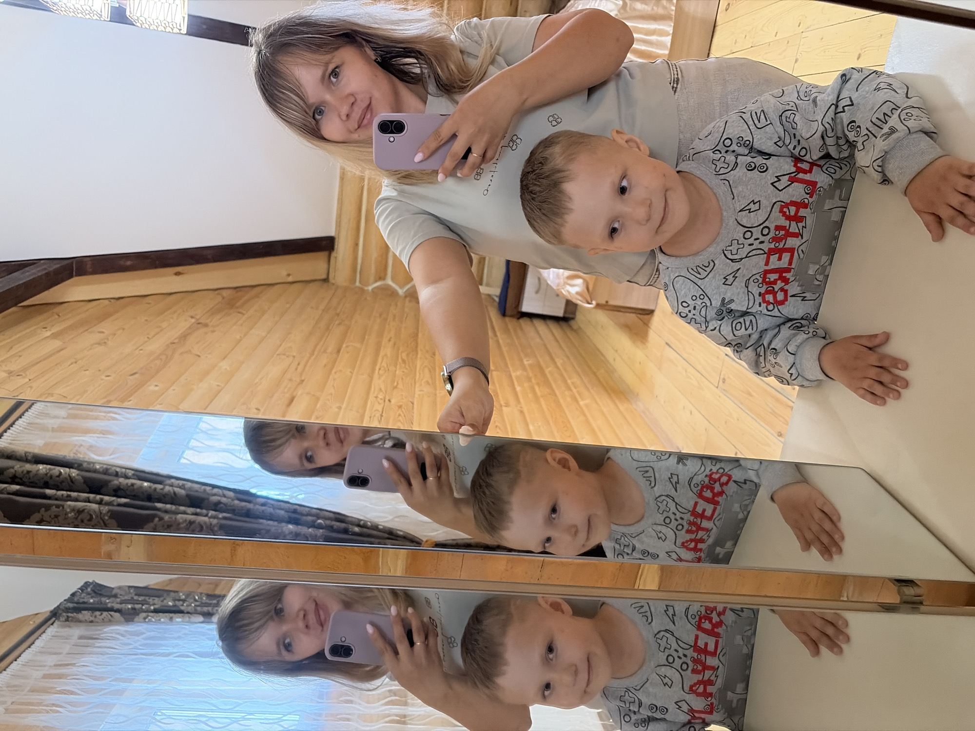An indoor mirror selfie captures a woman and a young boy, likely mother and son, in what appears to be a residential setting in Chaikovsky, Russia. The photograph is taken at an angle, emphasizing wooden floorboards and light-colored walls accented with dark wooden trim.
The woman, with fair skin and light brown hair, wears a light grey t-shirt and a silver-banded watch on her left wrist. She holds a light purple smartphone in her right hand, positioned to photograph their reflection in the mirror. A gold ring adorns her finger. She looks towards the camera with a subtle, content smile.
Beside her, a young boy with light brown hair and fair skin leans on a light-colored table. He is dressed in a grey long-sleeved shirt, decorated with grey game controller patterns and the distinct red text "PLAYERS" across the chest. He also looks towards the camera with a gentle smile.
The large, reflective surface, possibly a segmented mirror, captures multiple reflections of the subjects and parts of the room, including curtains or blinds visible in the background reflections. Both individuals appear calm and content. The scene conveys a relaxed, homey atmosphere, likely during daytime hours.