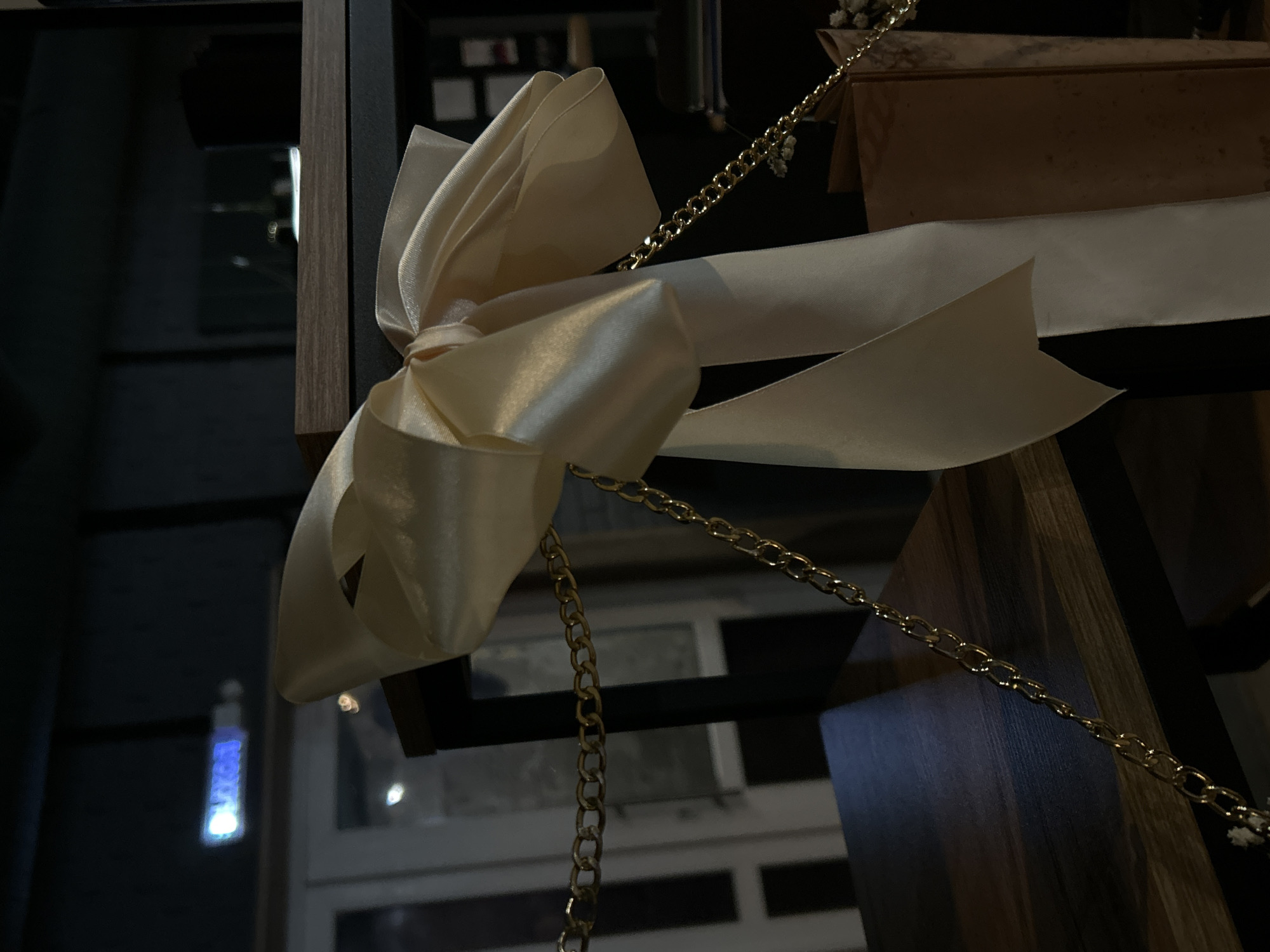 The image captures a close-up view of a decorative arrangement, primarily featuring a large, elegant light cream or off-white satin bow. The bow is prominently displayed, tied around a dark, slender structural element, which appears to be part of a shelf unit or display fixture made of dark wood and black metal or plastic.
A golden metal chain weaves through the composition, draping over and around the bow, and extending into the background. Small, delicate clusters of white flowers, resembling baby's breath, are visible attached to the chain near the top right. To the right of the bow, a light brown object, possibly a cardboard box or folded material, is partially visible.
The background is dark and features blurred reflections of lights, suggesting an indoor setting with subdued lighting, likely evening or night. One of the reflections, visible in the lower left portion of the image, is a blurred blue light with some indistinct white text, which is illegible. No people are present, and no specific activity is taking place; the scene is static and purely decorative. The overall impression is one of sophistication and understated elegance, part of an interior display in Chaikovsky, Russia.