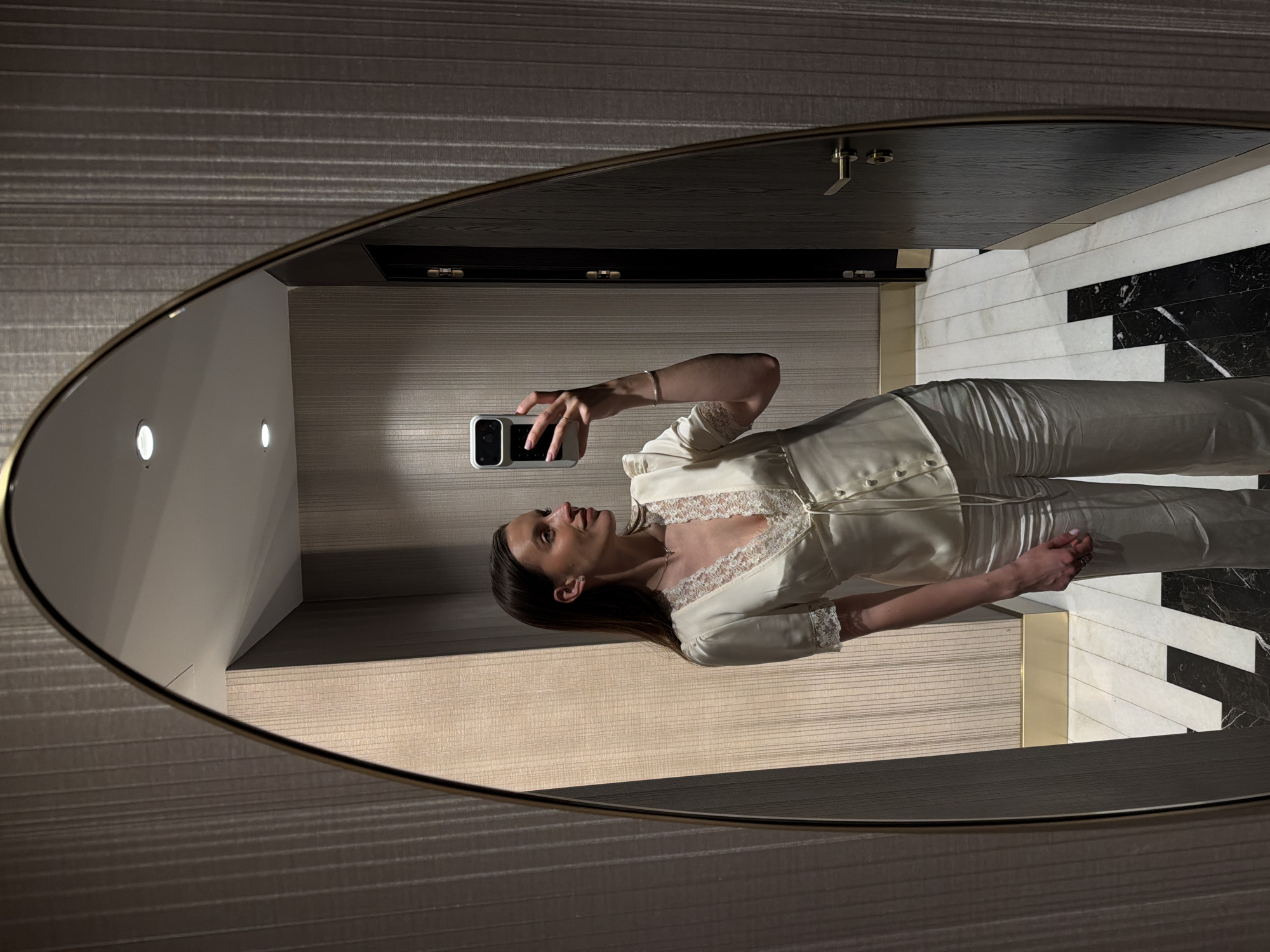 A woman is seen in a mirror taking a selfie with her phone. She is wearing a light-colored satin top with lace detailing and matching pants. The reflection shows a bathroom with black and white marble tiles. The wall behind her has a textured, light-colored wallpaper. There is a vanity mirror with a black top. The mirror itself has a thin golden frame. The image does not provide enough information to confirm the location in Budva, Montenegro.