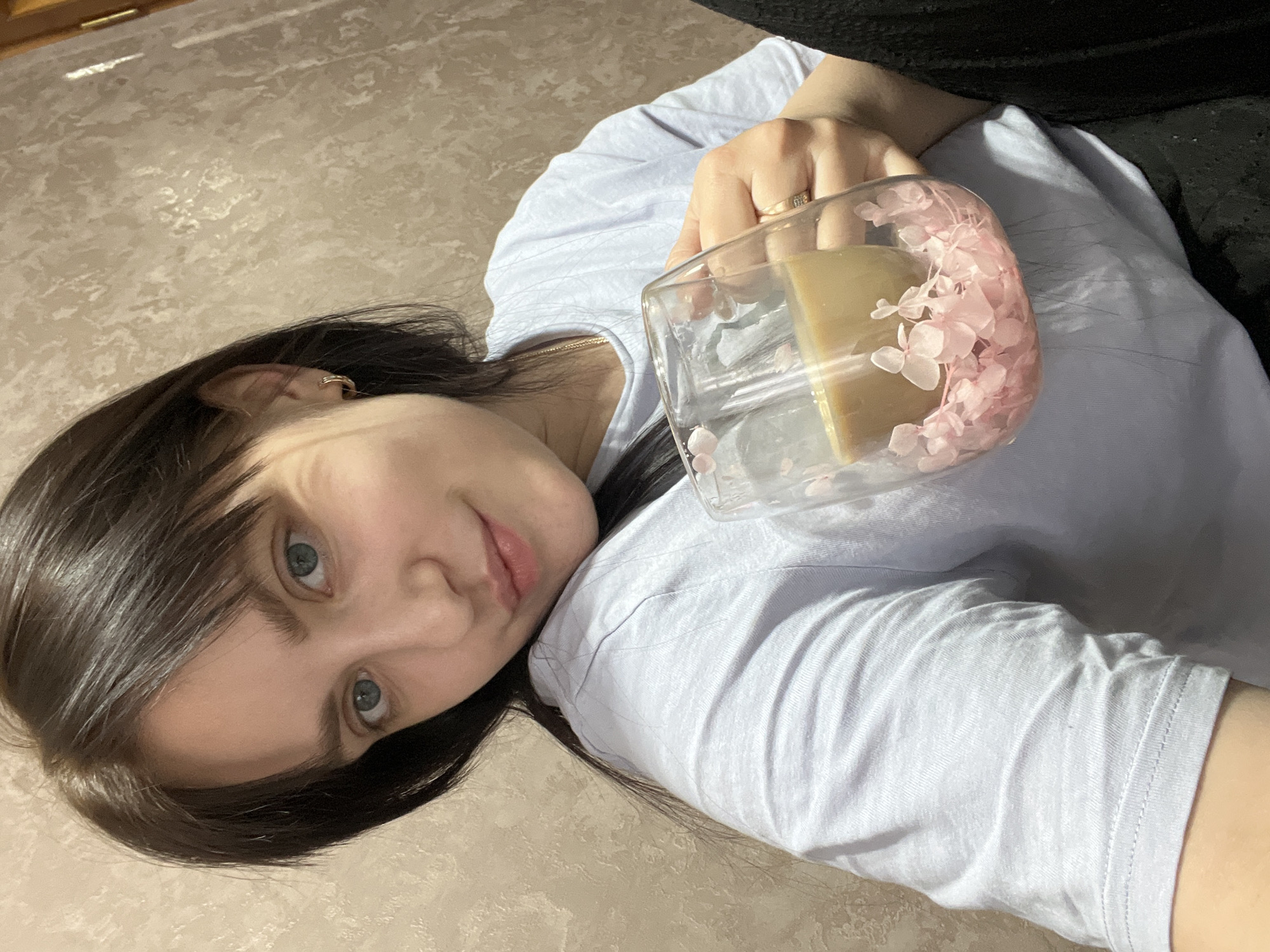 A young, fair-skinned woman with dark brown hair and blue eyes is lying down, holding a glass mug with pink floral decorations. The mug contains a light brown liquid, possibly coffee or tea. She is wearing a light blue t-shirt and what appears to be a black garment. The background is a textured beige surface, possibly a floor or a bedspread. The location is Марковский, Russia.