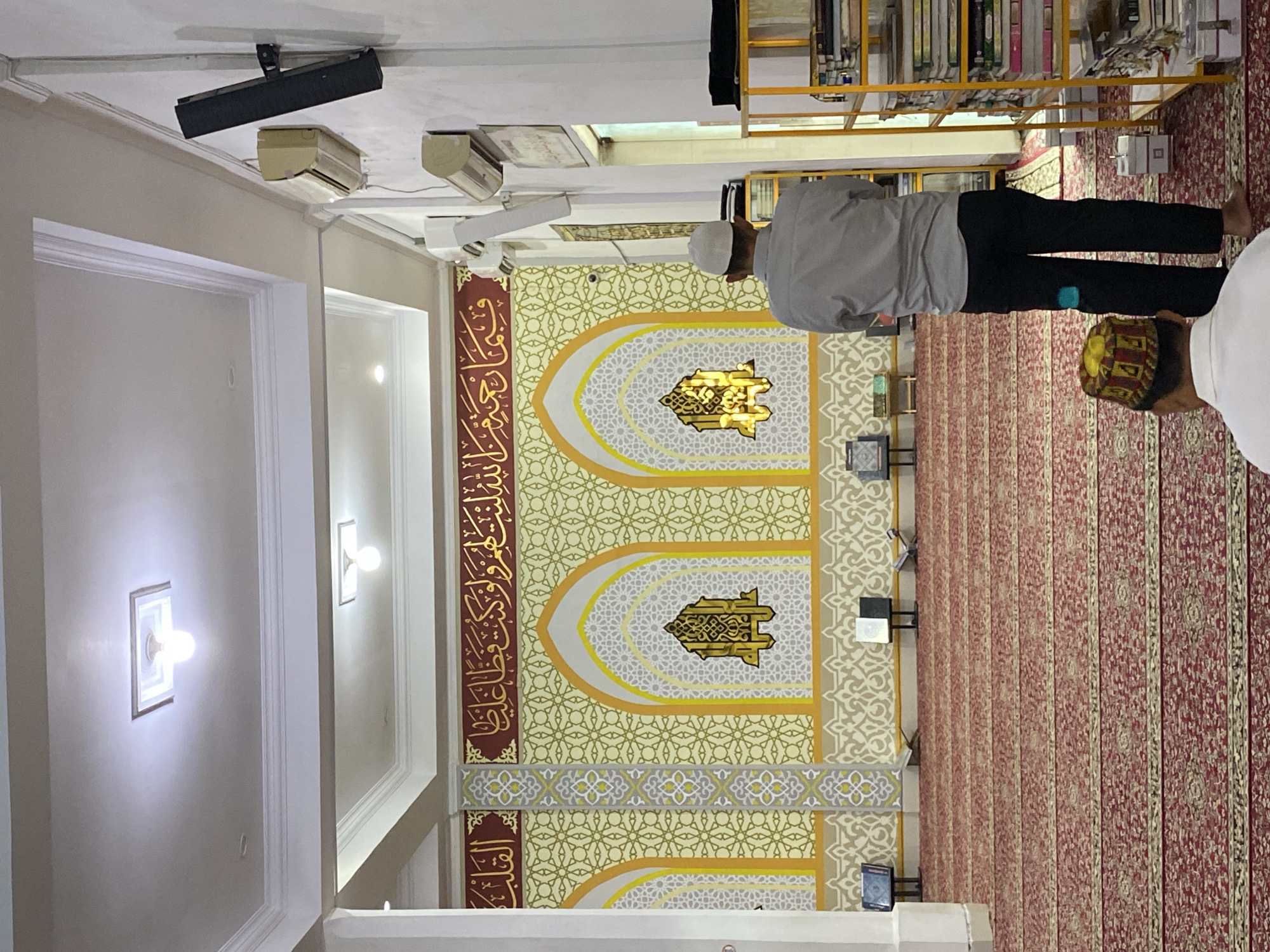 The image depicts the interior of a mosque, likely located in Lhokseumawe, Indonesia. Two men are present. In the foreground, viewed from behind, is a man wearing a white shirt and a colorful, patterned cap. Further back and also viewed from behind is another man wearing a light grey long-sleeved shirt and black pants, with a white cap. He appears to be standing. The mosque features ornate walls with yellow and white geometric patterns and Arabic calligraphy in gold against a red background. Arched decorative elements are visible on the wall. The floor is covered with a red carpet featuring intricate patterns. Shelving units filled with books and other items are positioned against the wall on the right side of the image. Air conditioning units and speakers are mounted on the wall. The ceiling has recessed lighting fixtures. Several book stands are placed on the carpeted floor in front of the decorative wall.