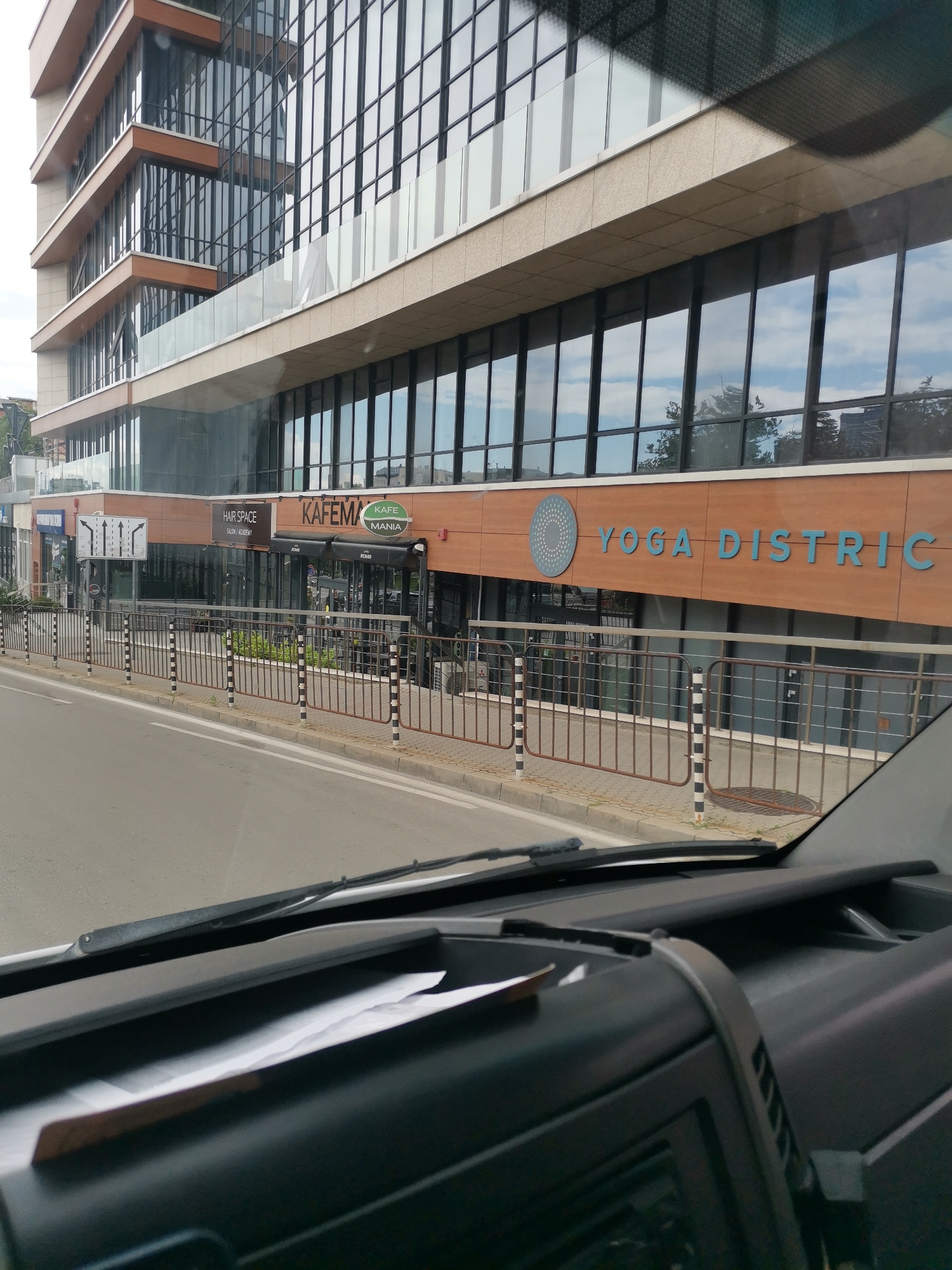 The image shows a street view in Sofia, Bulgaria, focusing on the facade of a modern building with large windows. On the ground floor, several businesses are visible. From left to right, a blue sign is partially visible, followed by a traffic sign showing directional arrows. Next, a sign for "HAIR SPACE SALON | ACADEMY" is displayed. To its right, a cafe named "KAFEMA" with a green circular logo is present, and further to the right, a sign reads "YOGA DISTRICT" with a circular blue graphic. A metal fence runs along the sidewalk in front of these establishments. The scene is captured from inside a vehicle, with the dashboard and windshield wipers visible in the foreground, suggesting the photo was taken while driving. The sky is partly cloudy.