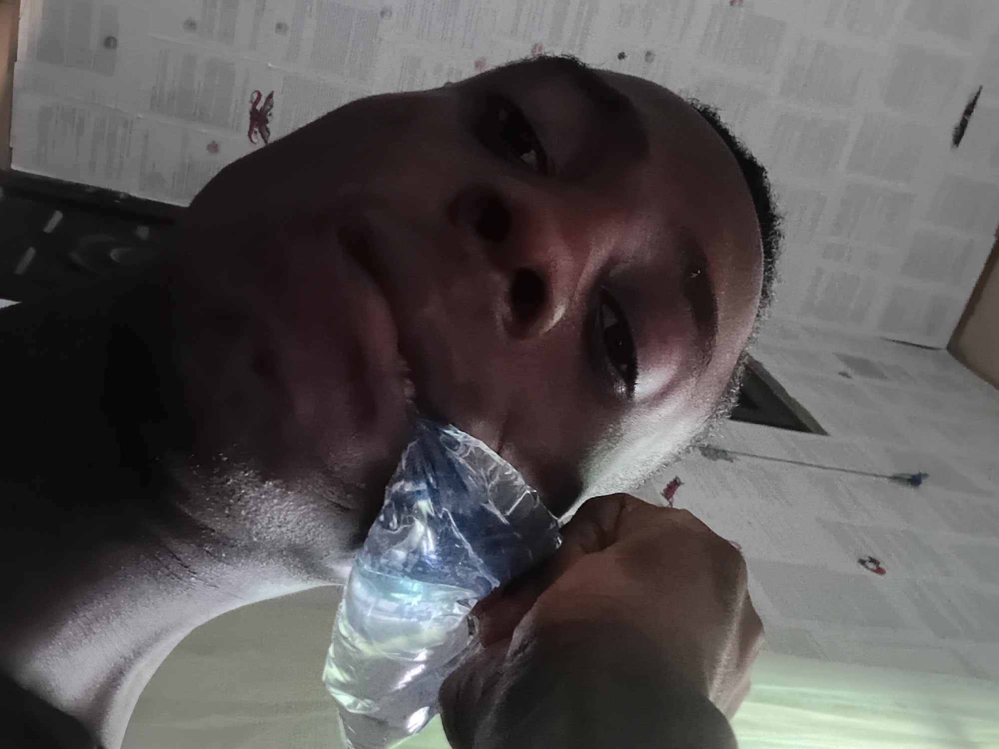 A person of African descent, likely male, is holding a plastic water bottle to their mouth. The person is facing the camera from a low angle. The background features a patterned wall, possibly wallpaper. The location in Akwete, Nigeria, cannot be definitively determined from the image.