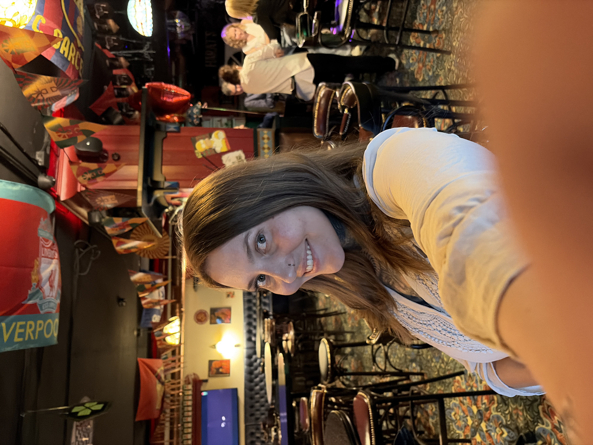 A woman with long brown hair and a nose ring smiles into the camera in a dimly lit pub. She is wearing a light-colored shirt with intricate embroidery around the neckline. The background shows a bar area with tables and chairs, some people standing and chatting. Decorative flags are hanging from the ceiling, and a television screen is visible on the far left. The floor is carpeted with a patterned design. The overall atmosphere suggests a casual and social gathering. The location is identified as a pub in Izhevsk, Russia.