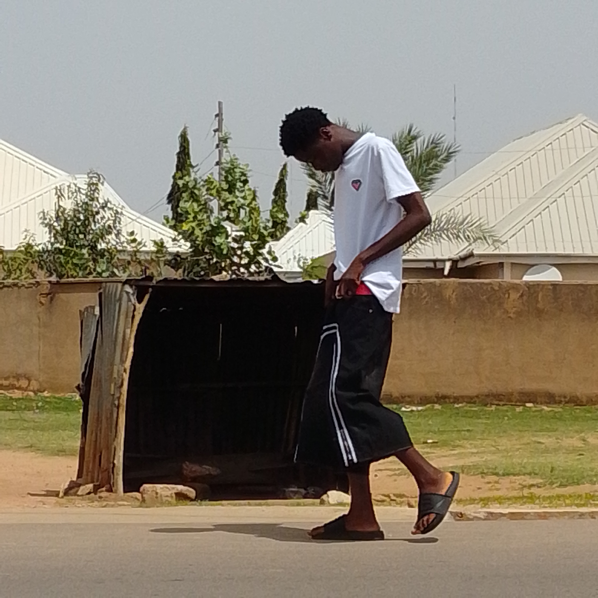 In Gombe, Nigeria, a young Black male wearing a white t-shirt with a pink heart logo and black pants with white stripes walks along a paved road. He is facing away from the camera, with his head tilted down. His right hand is in his pocket, and his left hand is by his side. He is wearing black sandals. In the background, a dilapidated wooden structure with a corrugated metal roof stands next to a tan-colored wall. Behind the wall are white-roofed buildings and lush green foliage, including palm trees. The sky is overcast.