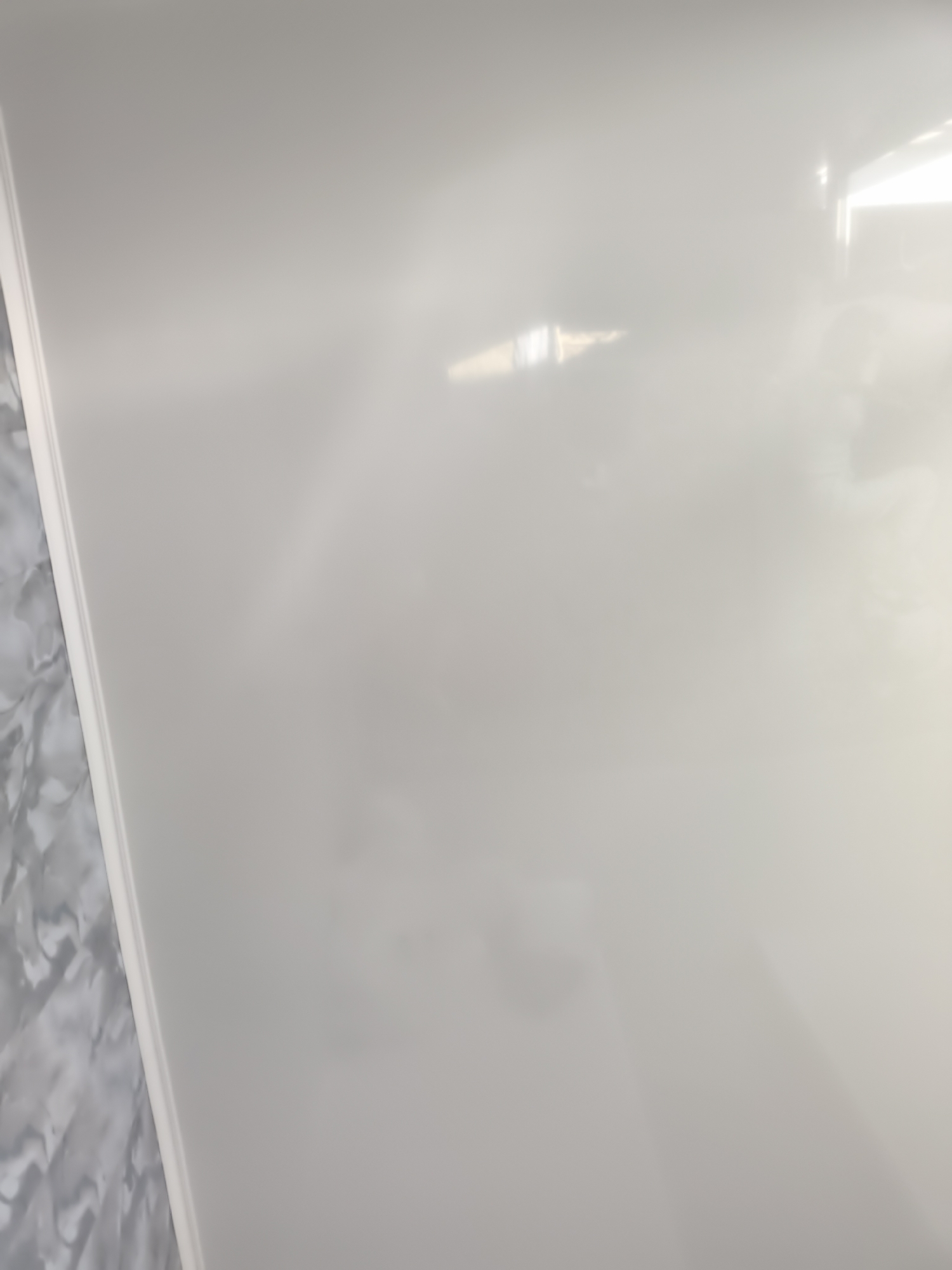 The image shows a glossy, white surface that appears to be a countertop or a table. The surface is highly reflective, with some blurred light and shadow patterns visible, suggesting an indoor environment with artificial lighting. On the left side of the image, a portion of a patterned surface, possibly marble or granite, is visible along the edge of the white surface. There are no people or discernible objects in the image beyond these surfaces. The image does not provide any specific visual cues to determine its location within Bishkek City, Kyrgyzstan.