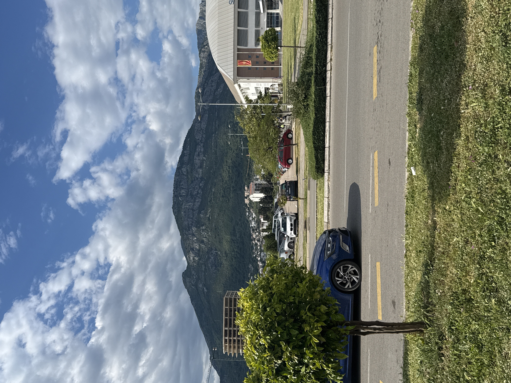 The image depicts a street scene in Bar, Montenegro. A large mountain looms in the background, partially obscured by clouds. A stadium or large building is visible at the base of the mountain. Along the right side of the image is a paved road with a yellow lane marking. Cars are parked along the roadside. A blue car is partially visible in the foreground. Grass and shrubbery border the road. The sky is blue with scattered clouds.
