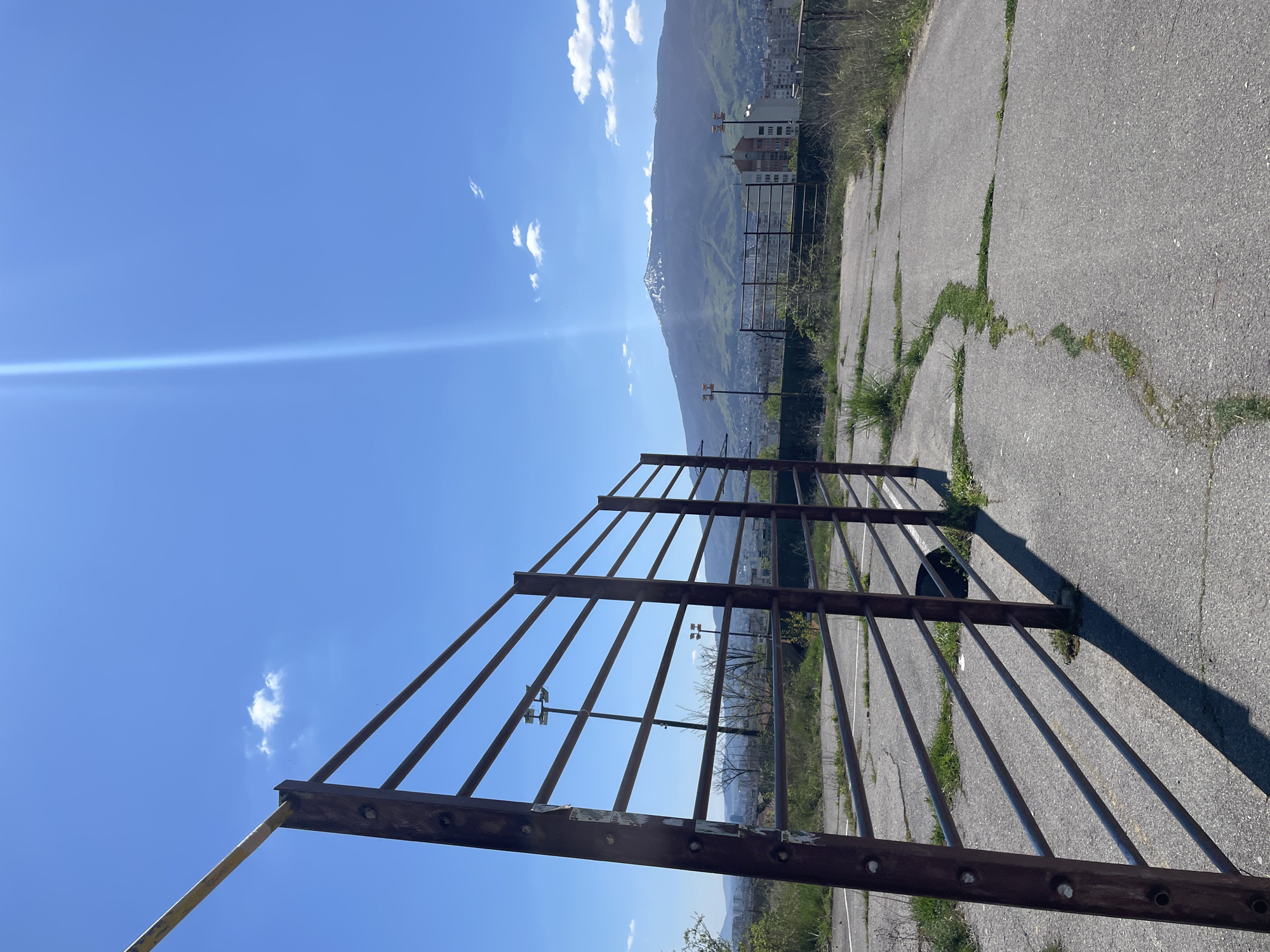 This media file captures an expansive outdoor scene in Sofia, Bulgaria, looking out from an elevated position over an urban area towards mountains under a bright, clear sky.
In the immediate foreground, a large, weathered metal fence or gate with numerous vertical bars stretches diagonally across the lower left portion of the image. The fence appears rusty brown, with some peeling paint or material on its horizontal supports. One yellow-colored bar extends from the fence towards the bottom left. The fence casts long, distinct shadows onto the ground.
The ground surface on the right side of the foreground and mid-ground is light gray asphalt, heavily cracked with green grass and weeds growing through the fissures, indicating an area of disuse or neglect.
Beyond the fence and asphalt, in the mid-ground, elements of an urban landscape are visible. Multi-story buildings, including a light-colored apartment-style block, are scattered among trees and greenery. Tall metal poles, possibly streetlights or floodlights, equipped with light fixtures, stand upright in this area.
The background is dominated by a majestic mountain range, stretching across the horizon. The mountains are primarily covered in green vegetation, with some higher sections showing rocky outcrops or patches of lighter-colored material, potentially snow.
The sky above is a vibrant, clear blue, adorned with a few scattered white cumulus clouds. A pronounced horizontal lens flare extends from the upper left, suggesting direct sunlight. The strong shadows and bright illumination indicate that the photograph was taken on a sunny day, likely during mid-morning or afternoon. There are no people visible, nor any discernible text. The scene presents a blend of urban development and natural landscape from a panoramic vantage point.