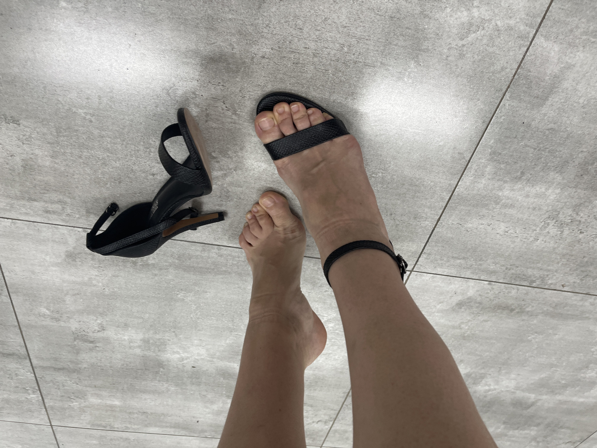 The image shows a person's legs and feet on a tiled floor. One foot is wearing a black sandal with an ankle strap, and the other foot is bare. A black high-heeled shoe is lying on the floor next to the bare foot. The person's legs are tanned. The tiled floor is a light gray color with darker gray grout lines. The image appears to be taken indoors. There is no indication of the location in the city of Afipskiy, Russia.