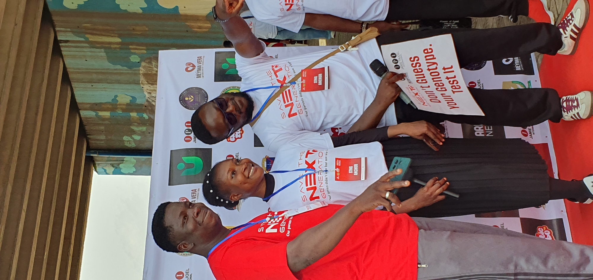 In Jos, Nigeria, three individuals stand on a red carpet in front of a white banner displaying logos. The person on the left, a dark-skinned male in a red t-shirt and grey track pants, smiles. To his right, a dark-skinned female with her hair in a bun, wearing a white t-shirt and black skirt, also smiles. A dark-skinned male with sunglasses, wearing a white t-shirt and dark pants, is on the far right, pointing towards something outside the frame. He holds a microphone and a white sign that reads, "Don't Guess Your Genotype. Test It." The banner behind them features logos of various organizations, including "SAVE THE NEXT GENERATION" and "MEYIWA VERA EMPOWERMENT FOUNDATION." The background suggests an outdoor event with a structure overhead.