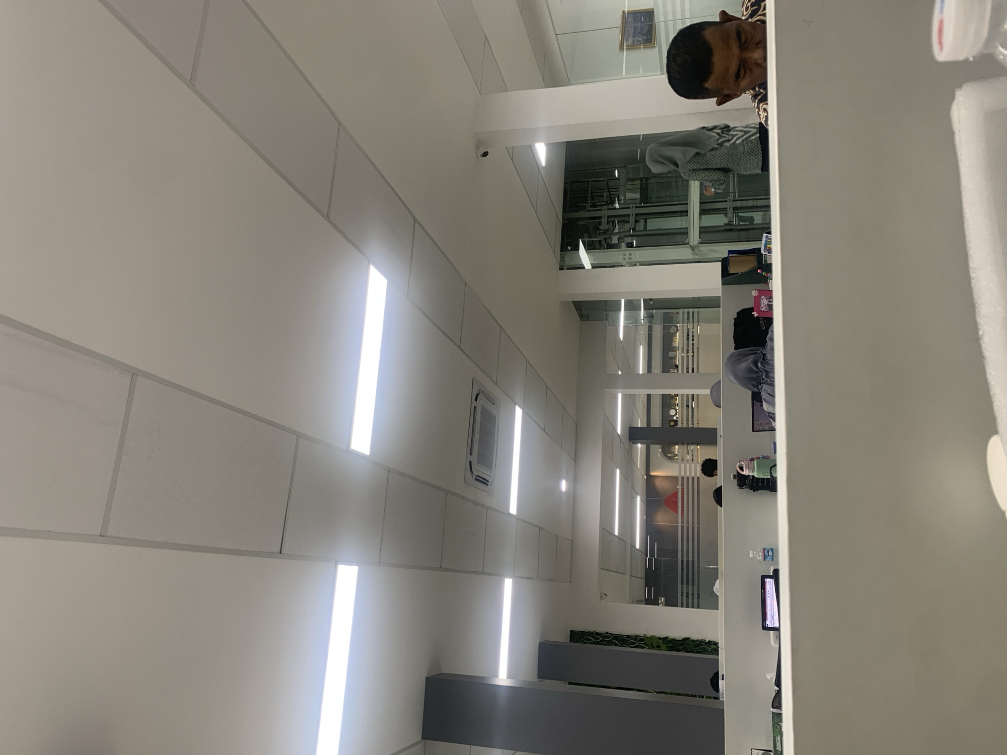 This image depicts an indoor scene, likely an office environment in Palembang, Indonesia. The ceiling is composed of white acoustic tiles with integrated rectangular fluorescent lighting fixtures. A central air conditioning unit is visible on the ceiling. The space appears to be divided by partitions, suggesting cubicles or workstations.
In the foreground, a man wearing a patterned shirt is partially visible, looking down. To his left, a person wearing a grey hijab is seated at a desk with a laptop. Further back, other individuals are discernible, some appearing to be working at desks with laptops and electronic devices. A woman in a patterned grey hijab is standing and looking towards the right. A security camera is mounted on a white pillar. The overall impression is that of a modern office setting.