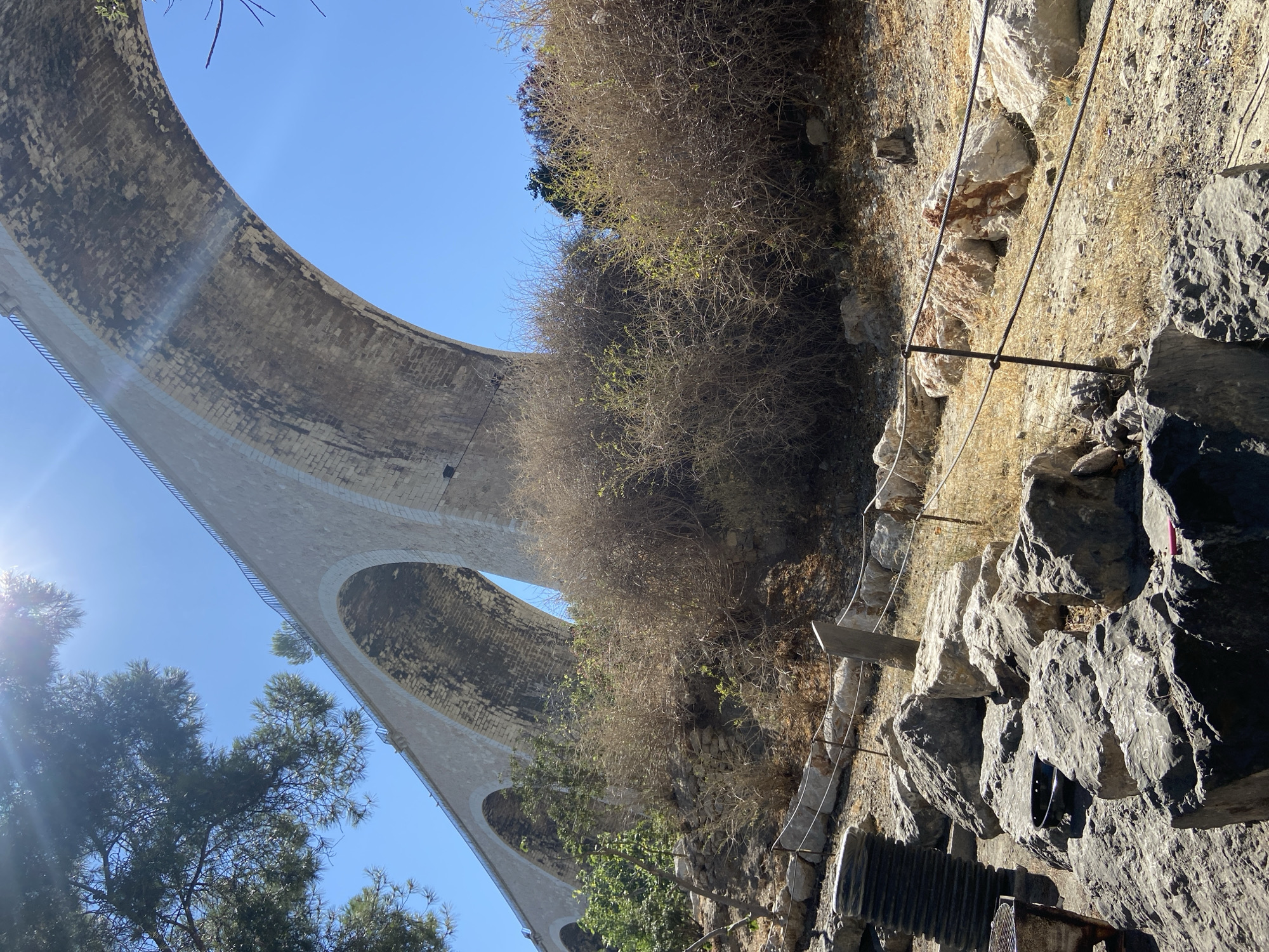 In an outdoor scene, a large, stone aqueduct with multiple arches spans across the frame, viewed from below. The aqueduct is constructed of light-colored stone blocks. The sky is clear and blue. In the foreground, the ground is covered with rocks, dry grass, and scrubby bushes. A metal fence runs along the middle ground, separating the rocky area from the vegetation. A large black cylindrical object, possibly a large pipe or a spool, stands to the left of the frame, next to a metal grill. Sunlight streams in from the upper left, casting lens flare and highlighting the top of the aqueduct.
