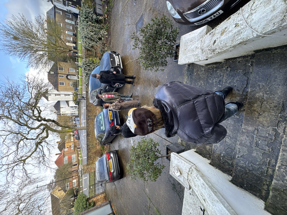 The image depicts a residential scene in Hampstead, London, United Kingdom, viewed from an elevated position looking down a set of worn stone steps into a wet, paved driveway.
In the immediate foreground, a person is seen from behind, standing on the stone steps. This individual wears a dark grey puffer jacket, blue jeans, and a brown baseball cap with "W 32" embroidered in gold lettering on the back. An empty white ceramic ashtray rests on a low stone balustrade to the left.
On the wet, dark grey paved surface below, five individuals are congregated among parked vehicles. One man, clad in a black overcoat and a dark hat, stands facing a group that includes a woman in a long brown coat and a man wearing a red and white striped sweater over a lighter shirt. Two additional individuals are partially visible within this gathering.
Four vehicles are present: a silver Jaguar sedan with license plate WP65 WXE, a dark blue sedan with license plate EO07 JGP, a black Jaguar SUV with license plate WM67 ACF, and the front right portion of a brown Lexus with license plate NB05 XAU.
The background shows a street lined by a low brick wall and dense green hedges. Across the street, a variety of residential buildings are visible, characterized by traditional architecture including red-roofed brick houses, a distinct white building featuring a turreted roof, and several terraced houses. Bare deciduous trees are silhouetted against an overcast sky that shows patches of blue. Distant pedestrians are discernible on the street.