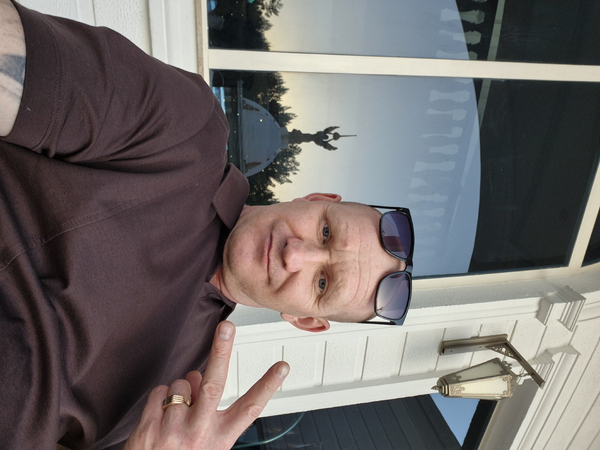 A middle-aged Caucasian man is pictured outdoors, wearing a brown collared shirt and sunglasses perched on his head. He is giving a "peace" sign with his right hand, on which a gold ring is visible. Behind him is a window reflecting an image of what appears to be a statue, specifically the Atatürk Monument in Alanya, Turkey. The man is standing near a white-painted building, which includes a decorative outdoor light fixture and siding. The scene appears to be taken during the day.