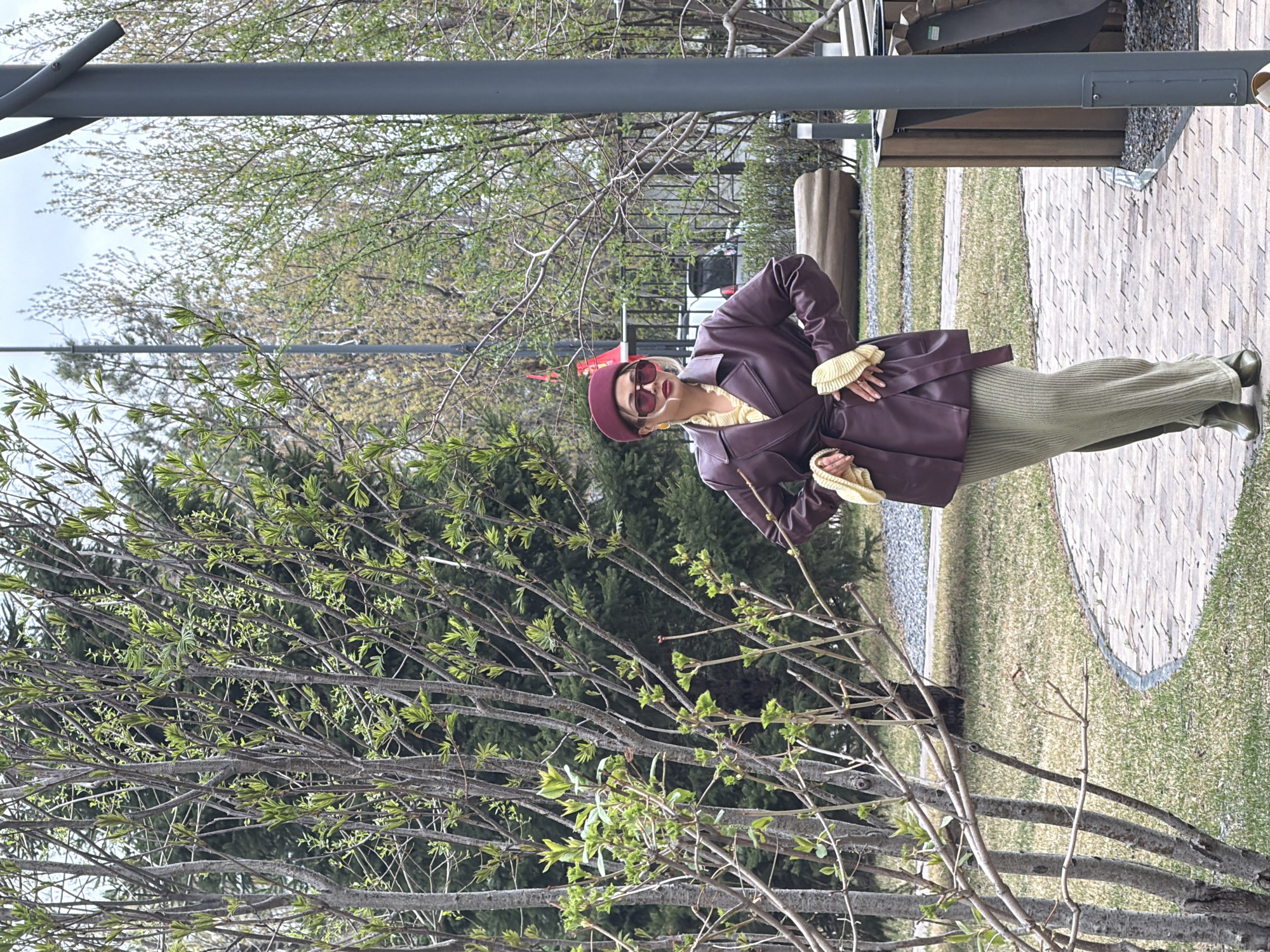 A woman in a burgundy hat, matching jacket, and green dress stands in a park. She wears sunglasses and holds a light-colored object in her hands. A tree with budding leaves is in the foreground. A gray metal structure is visible in the background, along with a wooden bench and a pathway made of light-colored materials. The location appears to be in Krasnoyarsk, Russia, based on details from other similar images.