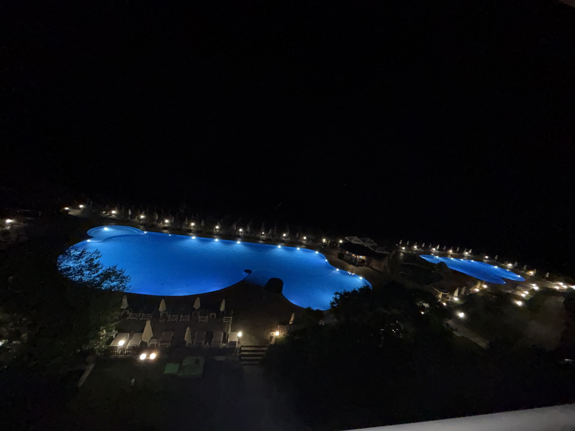 An elevated, wide-angle, night shot shows two illuminated swimming pools at a resort in Varna, Bulgaria. The larger pool, in the foreground, is a brilliant blue and appears to be an infinity pool. The smaller pool, in the background, is also illuminated and appears to be a standard shape. Both pools are surrounded by lounge chairs and umbrellas, most of which are not in use. Lights are visible along the edge of the pools and in the surrounding trees. The scene is dark, with the exception of the lights from the pool and surrounding areas.
