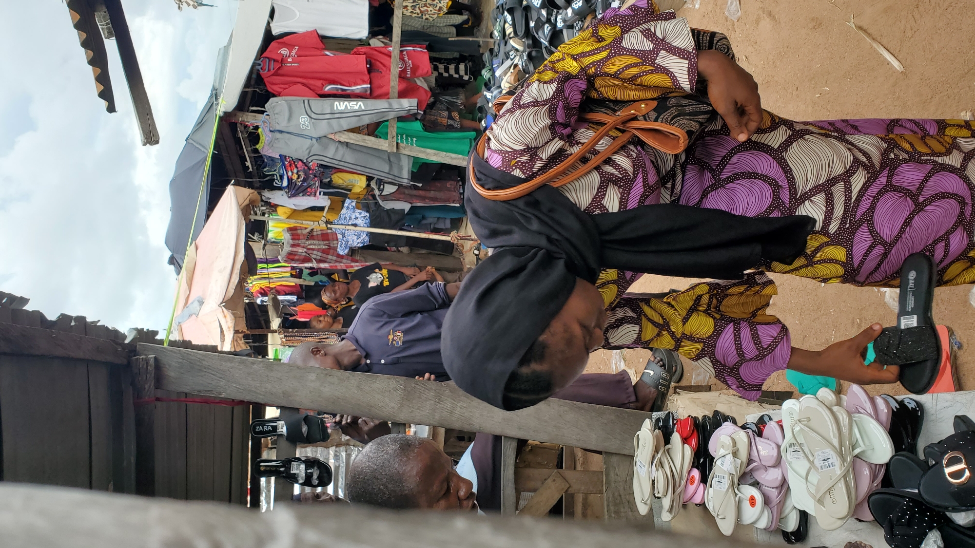 The image depicts an outdoor market scene in Jalingo, Nigeria, where people are browsing and shopping for footwear. In the foreground, a woman wearing a black headscarf and a colorful patterned dress is examining a black slipper. She is holding the slipper in her right hand and appears to be considering a purchase. To her left, a stack of assorted sandals and flip-flops is neatly arranged on a surface, suggesting a vendor's stall.
Further back, several other individuals are visible. A man in a blue shirt is looking towards the side, while another man is partially obscured by a wooden pillar. In the background, racks of clothing, including shirts and pants, are displayed under makeshift shelters, indicating a bustling marketplace. The ground appears to be sandy or dusty, typical of outdoor markets in the region. The sky above is overcast with clouds. The overall impression is one of everyday commerce and local community life in Jalingo.