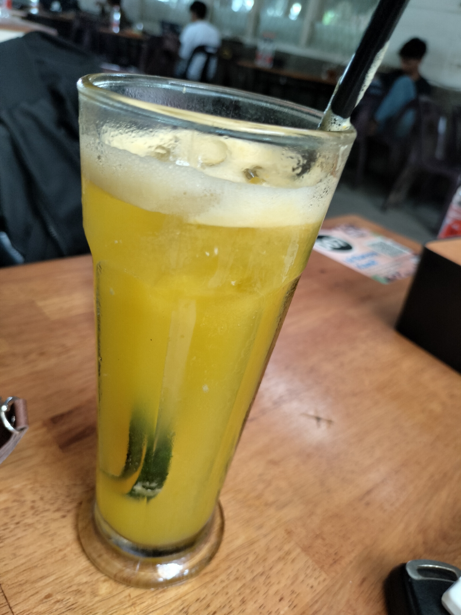 A tall glass of bright yellow juice with a frothy top sits on a wooden table. Two green wedges are submerged in the juice, and a black straw is inserted into the glass. The glass is beaded with condensation. In the background, blurred figures of people are visible in what appears to be an indoor setting, possibly a cafe or restaurant. A black jacket is draped over the back of a chair to the left of the glass. The table is made of light-colored wood. The image was taken in Langsa, Indonesia.