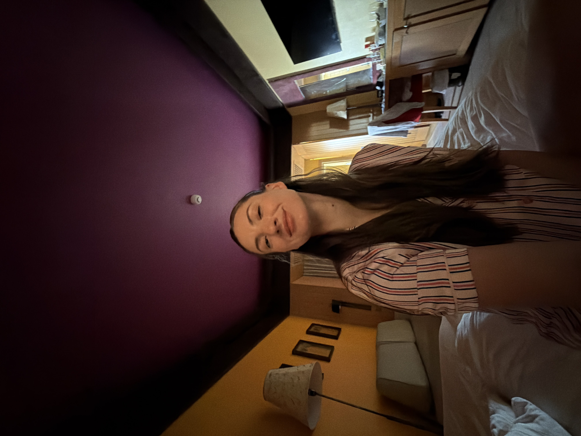 A woman is taking a selfie in a bedroom. She is wearing striped pajamas and has long brown hair. She is smiling at the camera. The room is decorated in a bold style with a purple ceiling and yellow walls. There is a bed in the foreground, and a desk with a television in the background. The image is taken from a low angle, looking up at the woman and the ceiling. This scene is in a hotel room in Dahab, Egypt.