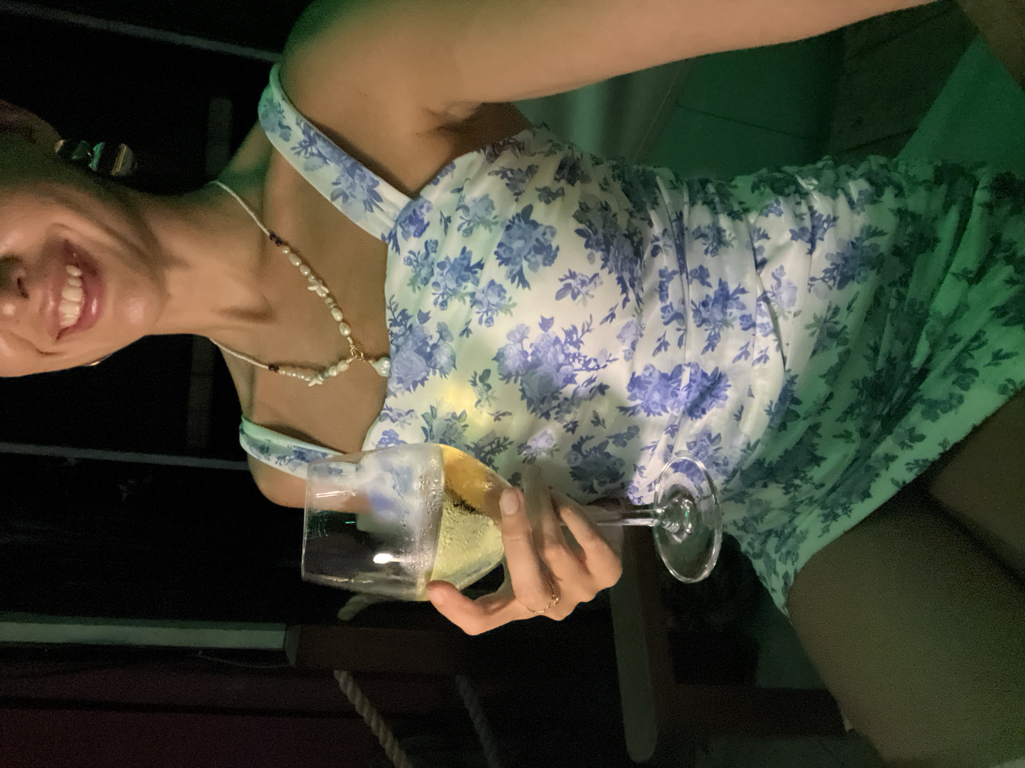 The image, taken in North Kuta, Indonesia, features a person, likely female, captured from the chest up in a diagonal orientation. She is smiling broadly, revealing her teeth, and appears joyful. Around her neck, she wears a pearl necklace interspersed with small, distinct charms and darker beads. She is dressed in a light-colored, possibly white or pale yellow, dress adorned with a blue floral pattern of various flowers and leaves, featuring a halter-style strap.
In her right hand, she holds a stemmed glass filled with a light yellow, effervescent beverage, likely champagne or sparkling wine. A delicate ring is visible on her ring finger. The setting is dimly lit, suggesting it is nighttime, with a prominent green light source casting a hue on the background, parts of her dress, and her skin. The background is predominantly dark, showing hints of dark wooden structures and a rope detail in the lower left, possibly indicating an outdoor or bar environment. No visible text is present in the media.