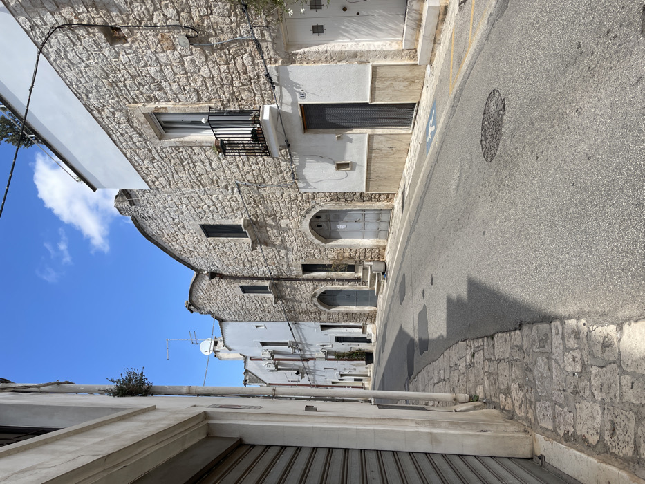 The image depicts a deserted street in Ceglie Messapica, Italy, under a clear blue sky with sparse white clouds. The asphalt road, appearing to ascend slightly, is flanked by buildings on both sides.
On the left, the immediate foreground features a light-colored building with a large, closed metal roll-down security shutter covering its ground-level entrance. Further along this side, multiple white-painted structures with visible air conditioning units and various windows and doors line the street. Rooftops in the distance display television antennas and a satellite dish.
The right side of the street is lined with buildings primarily constructed of light-colored stone with white upper sections and trim. A prominent building features a small, black wrought-iron balcony on the first floor, containing a potted plant. Below the balcony, the ground floor has a white facade with a light brown wainscoting, an address number "15," and a doorway protected by a metal chain-link screen. Adjacent to this, a white wooden door with small square window inserts is visible. Further along this side, multiple buildings feature distinctive arched doorways, some with small sets of stone steps leading up to them. These doors are typically gray or metallic in appearance. Windows on these stone buildings are dark, suggesting closed shutters.
The street surface includes a manhole cover near the center and a blue disabled parking symbol painted on the right sidewalk. Yellow painted lines are visible on the curb near the right sidewalk. A narrow, uneven cobblestone sidewalk borders the left side of the asphalt road. Strong sunlight from an elevated source casts distinct shadows of the buildings and their features onto the street and sidewalks. No individuals or vehicles are present. Minimal vegetation, including a small plant growing from the side of the leftmost building and the plant on the balcony, is visible.