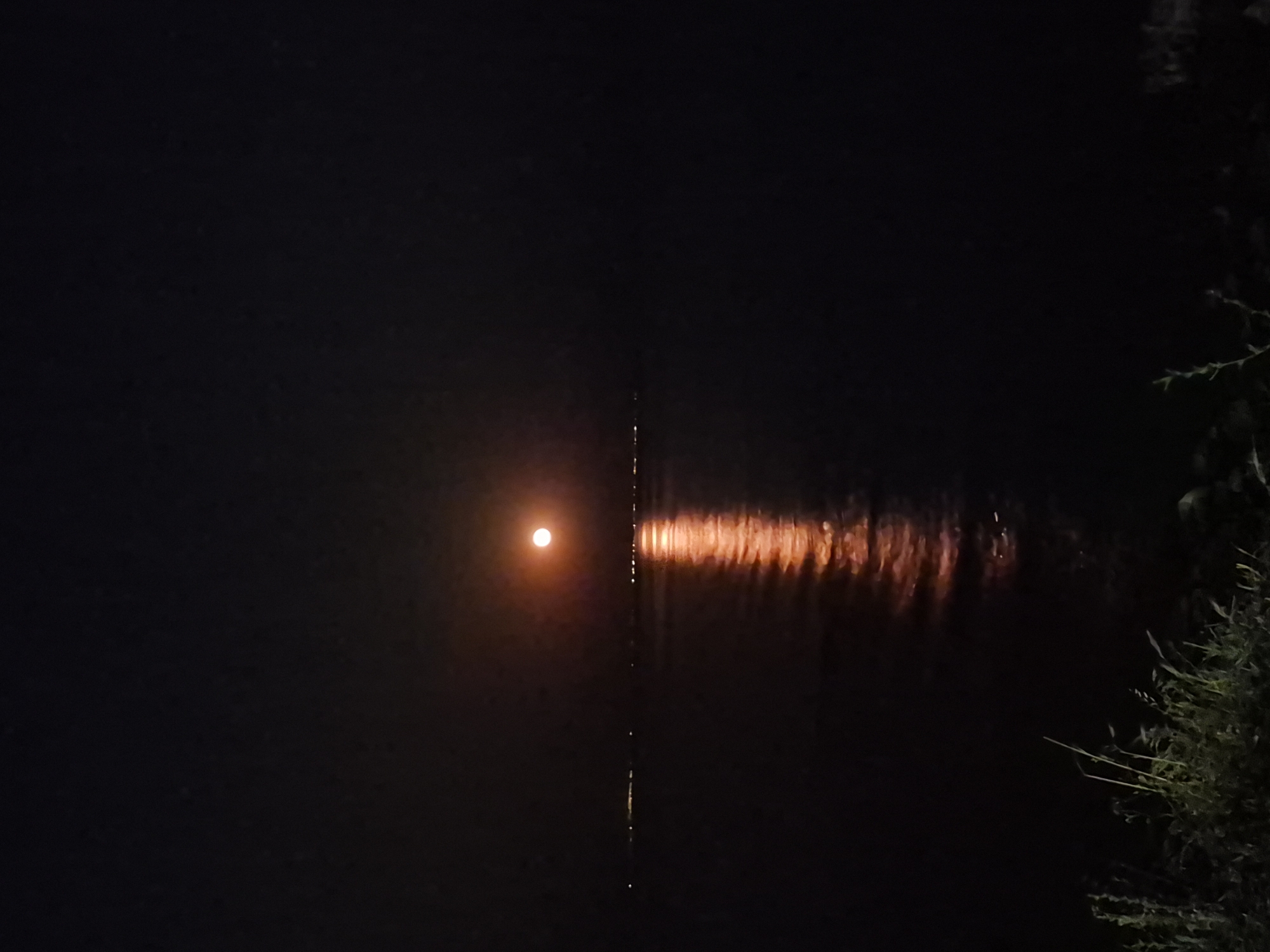 A full moon, appearing orange, is positioned in the upper center of a dark sky. The moonlight casts a shimmering, orange reflection onto the surface of a body of water, which occupies the lower half of the image. A faint line of distant lights, possibly from a town or city, is visible on the horizon across the water. In the foreground, dark vegetation, possibly grass and reeds, is visible along the bottom edge of the frame. The overall scene depicts a nighttime view of a moonlit waterfront in Makri, Greece.