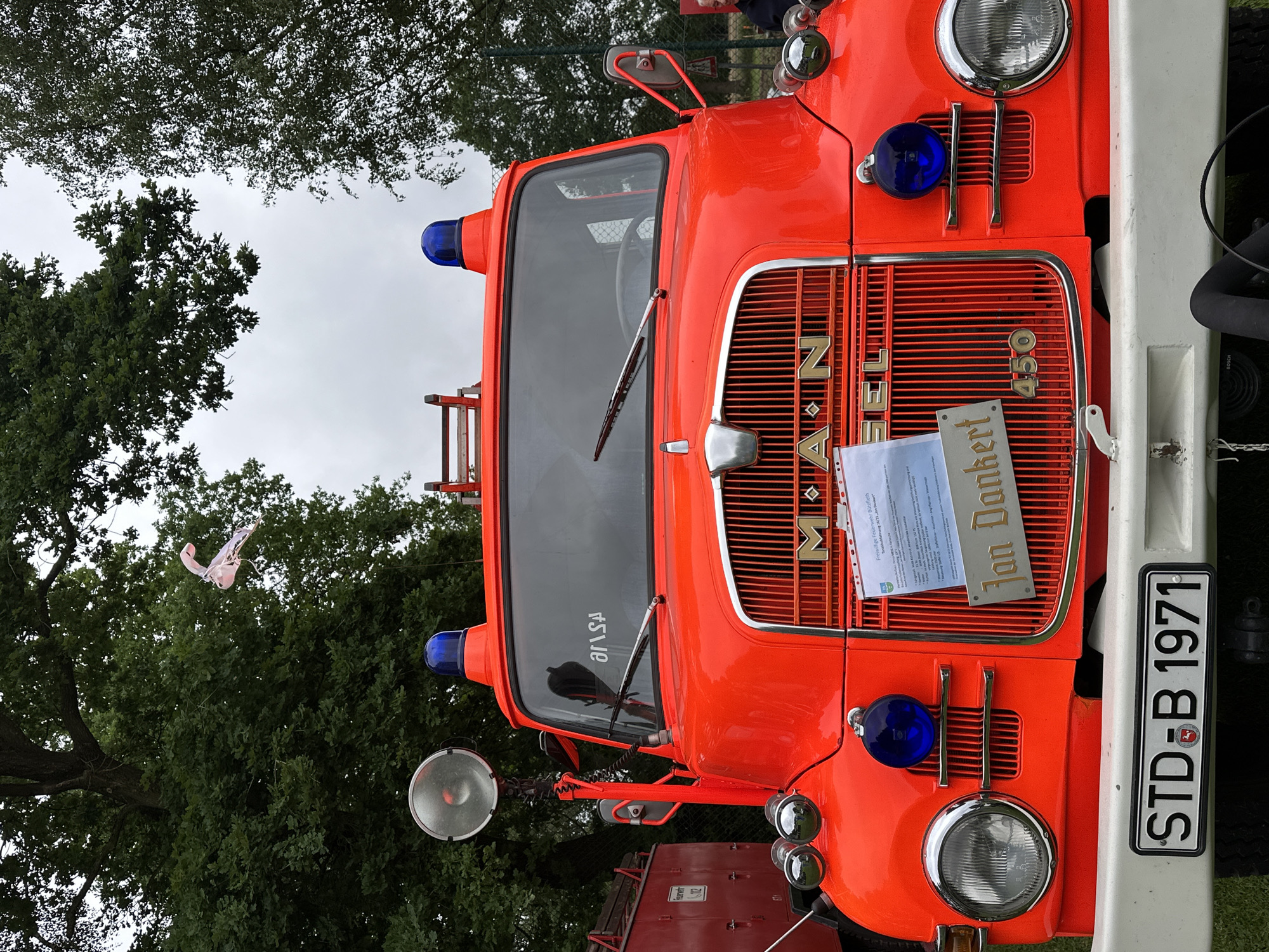A vintage bright red MAN DIESEL 450 fire truck is centrally positioned, facing the viewer. The truck features a chrome grille with the "MAN" and "DIESEL" logos prominently displayed, along with the number "450" on the lower right of the grille. Two round blue emergency lights are mounted on the roof, with a large chrome spotlight or siren visible on a pole to the left of the windshield. Below the grille, two large round headlights are present, flanked by smaller, round blue lights. A white front bumper runs across the base of the vehicle.
A white document, likely providing information about the vehicle, is attached to the grille on the left side, and a golden plaque reading "Jan Dankert" is affixed to the grille's lower-center section. The license plate "STD∙B 1971" is prominently displayed on the front bumper. The windshield reflects part of the surroundings, including a partially visible "arsA" text, reversed, suggesting a reflection from an external source or a sticker inside the cabin.
In the background, a dense canopy of green trees fills the upper portion of the frame, indicating an outdoor setting. The sky is overcast and grey. To the far left, a portion of another red vehicle, resembling a fire truck, is visible, displaying a white sign with "Feuerwehr 112". A light-colored, possibly white or translucent, irregularly shaped object, potentially a deflated balloon or plastic debris, is floating in the upper-mid section of the image, suspended against the trees. A faint mesh fence and distant trees are visible on the right side. The scene is static, depicting the fire truck parked outdoors, possibly at an exhibition or event in Deinste, Germany.