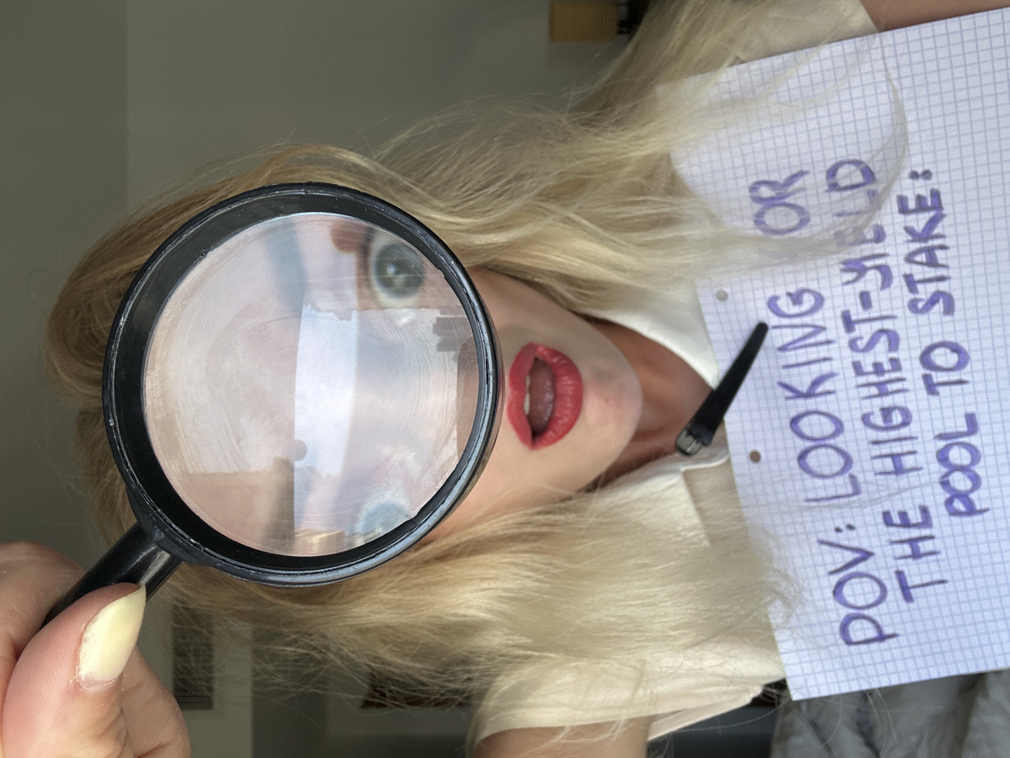 The image shows a close-up of a woman's face, partially obscured by a magnifying glass she is holding. She has blonde hair and is wearing bright red lipstick. The magnifying glass is held over her eyes, which are visible through the lens and appear enlarged. She is holding a piece of graph paper with purple handwritten text that reads: "POV: LOOKING OR THE HIGHEST-YIELD POOL TO STAKE:". The background appears to be an indoor setting, possibly a room in a hotel or apartment, with a muted color palette. There is no discernible connection to the city of Petah Tikva, Israel.