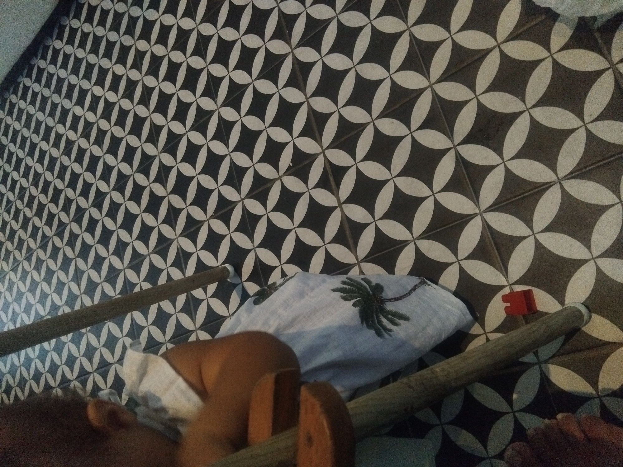 The image shows a tiled floor with a black and white floral pattern, and part of a human body, possibly a torso and leg. There's a wooden frame or structure partially visible, perhaps a low bed or seat, with a cushion or pillow patterned with a palm tree design placed on it. A red plastic object lies on the tiles near the edge of the frame. The lighting appears to be somewhat dim or indirect. While the location is given as North Kuta, Indonesia, there are no location-specific identifiers visible in the image itself to confirm this.