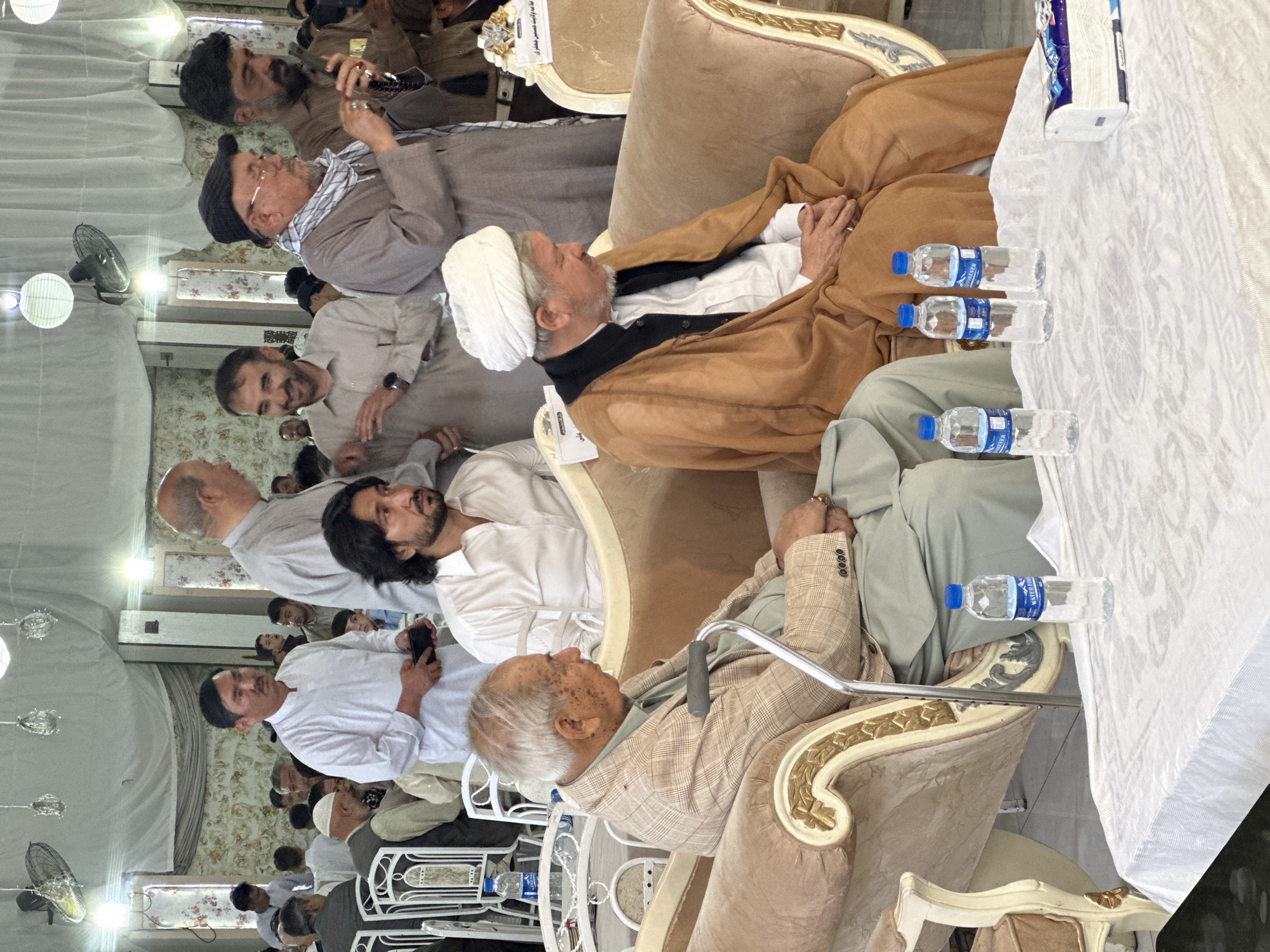 The image shows a gathering of men, possibly in Quetta City Tehsil, Pakistan. Several men are seated on ornate chairs or couches. Some individuals are wearing traditional clothing, including head coverings. There are water bottles and tissues on a table with a white tablecloth. Some people are using mobile phones. The environment appears to be indoors, with chandeliers and wallpaper in the background.