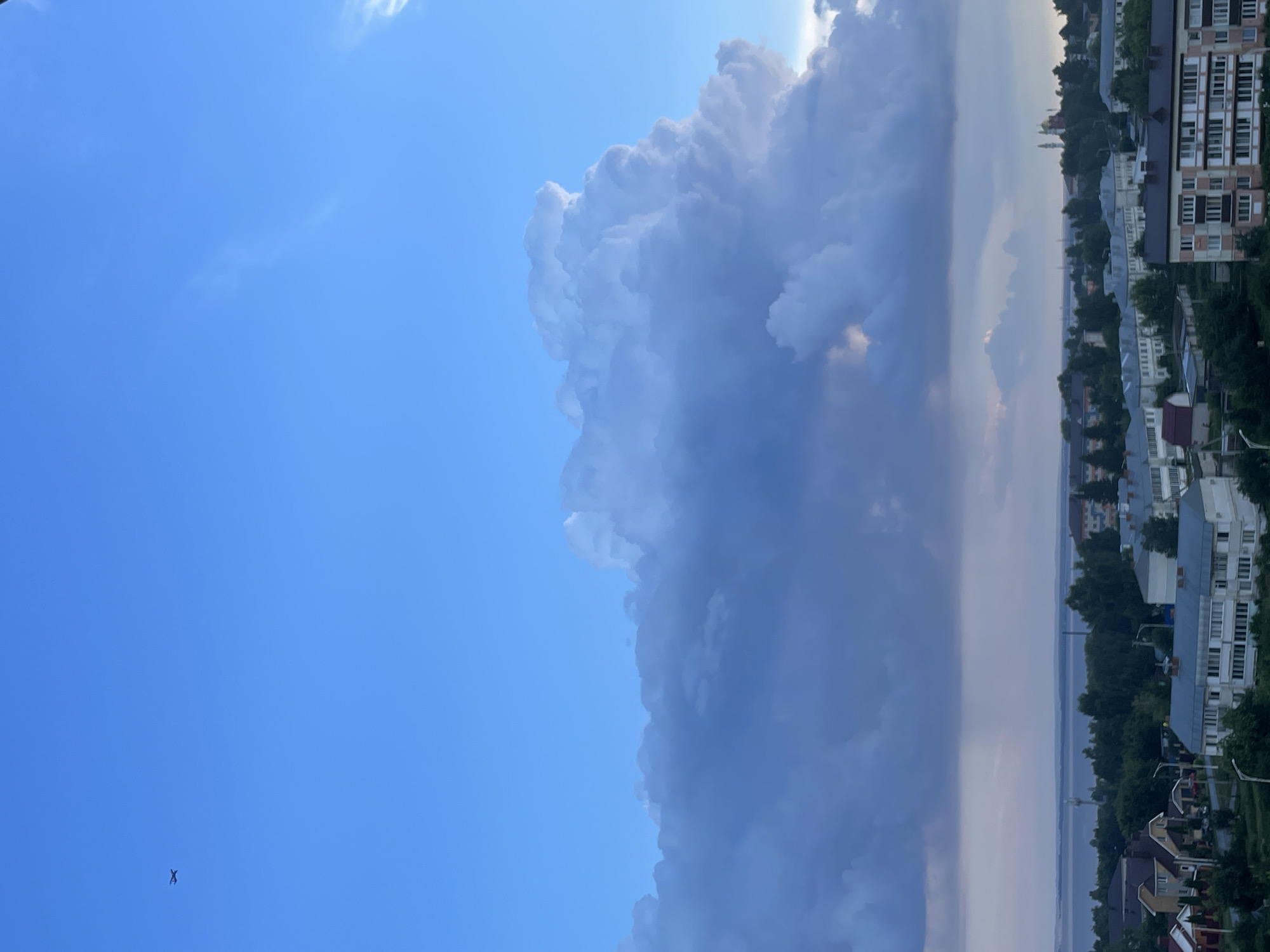 The image captures a view of the city of Laishevo, Russia, under a vast, partly cloudy sky. In the upper left quadrant of the frame, a small, dark airplane is visible against the clear blue sky. The dominant feature of the sky is a large, towering cumulus cloud formation, casting shadows over the landscape below. The city itself is nestled between a body of water, likely a reservoir or river, and the dense greenery of trees. Buildings of various sizes and architectural styles are visible, including residential houses and apartment blocks. The overall impression is one of a serene, possibly late afternoon or early evening scene, with the dramatic cloud formation adding a sense of dynamism to the otherwise peaceful panorama.