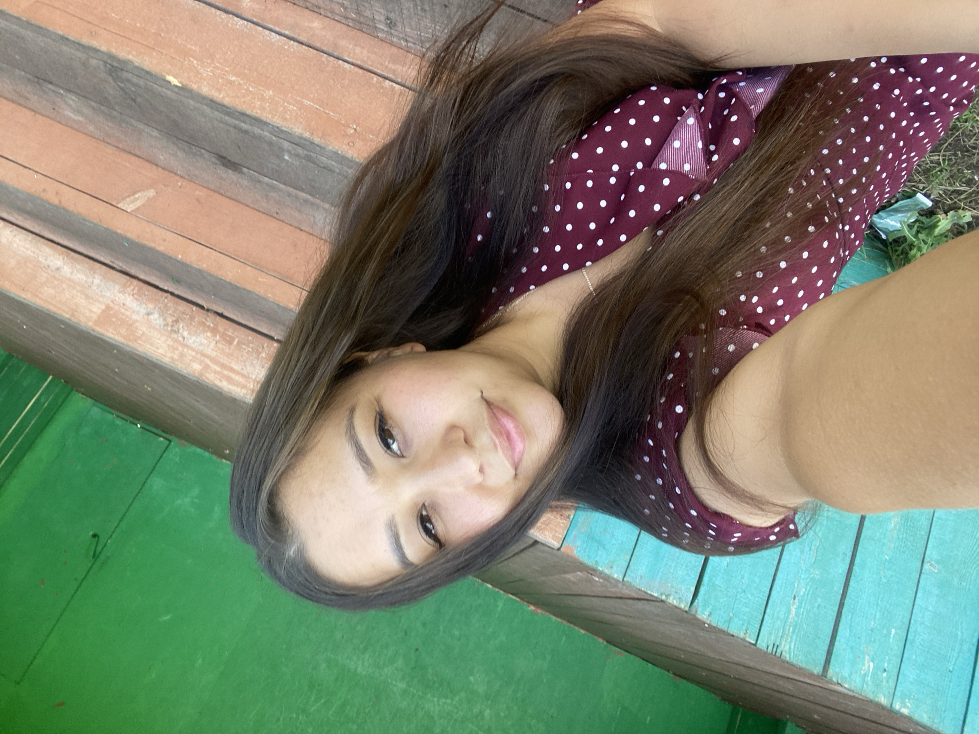 A young woman is taking a selfie outdoors. She is wearing a maroon dress with white polka dots and a thin silver necklace. Her long, dark brown hair is down and falls around her shoulders. She is smiling at the camera. The background shows a green wooden wall and a set of wooden steps. The location is identified as Maloarslangulovo, Russia.