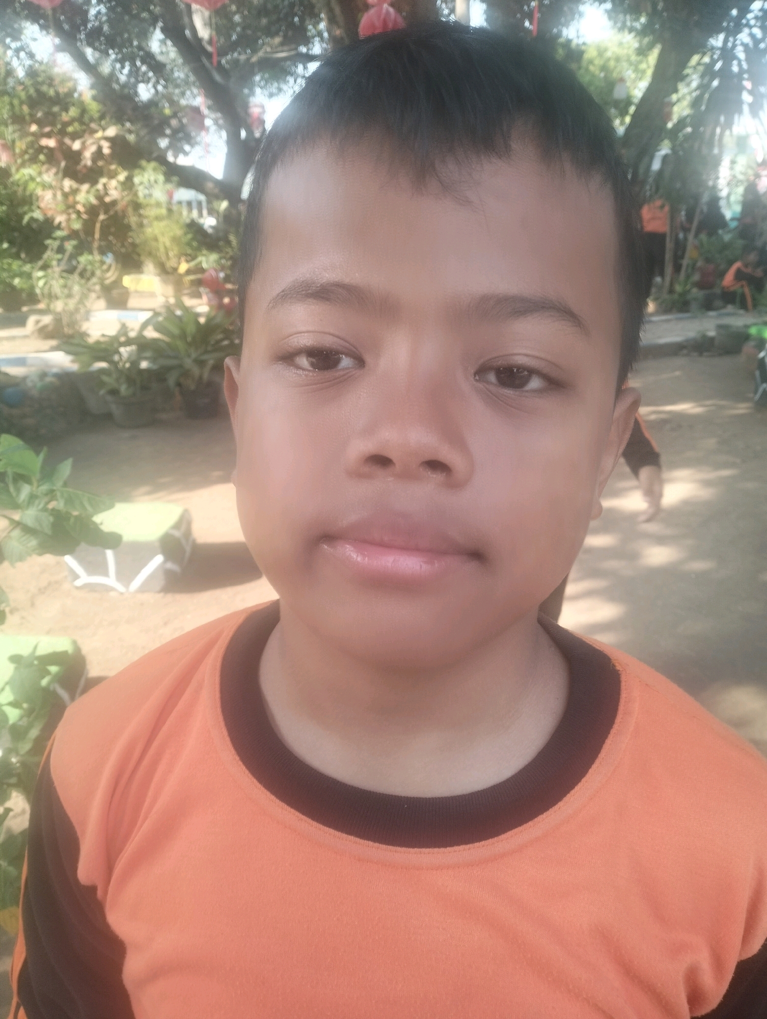 The image is a close-up portrait of a young boy wearing an orange shirt with black sleeves and collar. He has short dark hair and is looking directly at the camera. The background appears to be outdoors, with trees, plants, and what looks like a dirt path. There are hints of other people and structures in the blurred background. The setting suggests a schoolyard or park environment. The lighting indicates it is daytime, likely sunny due to the shadows. There is no specific indication of the city of Blabakan, Indonesia, within the image itself.