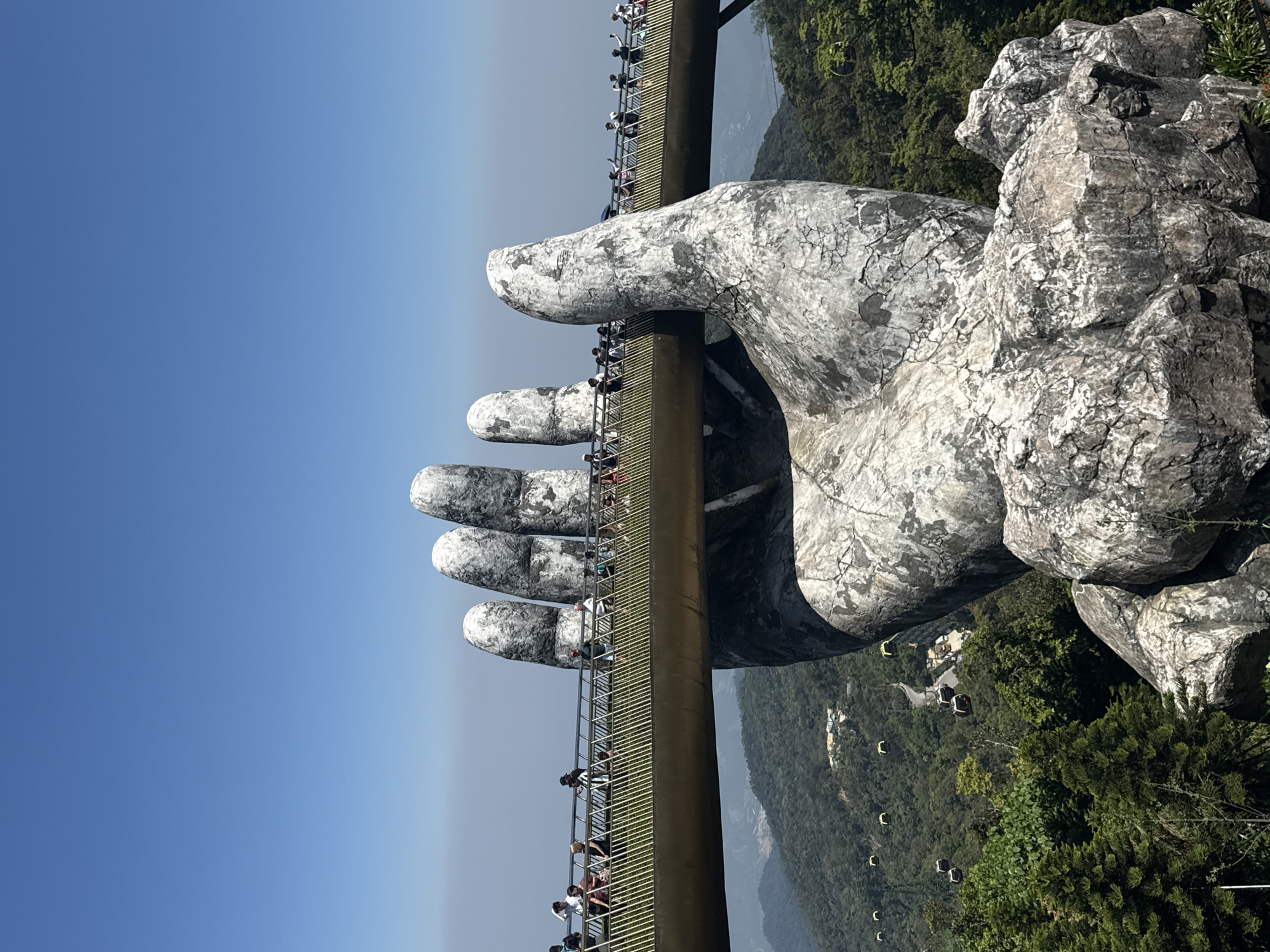 The image displays the Golden Bridge at Bà Nà Hills in Đà Nẵng, Vietnam. The bridge is supported by two giant, sculpted stone hands. People walk along the bridge, which spans the landscape. The backdrop consists of forested mountains and a clear blue sky. In the valley, there are cable cars.