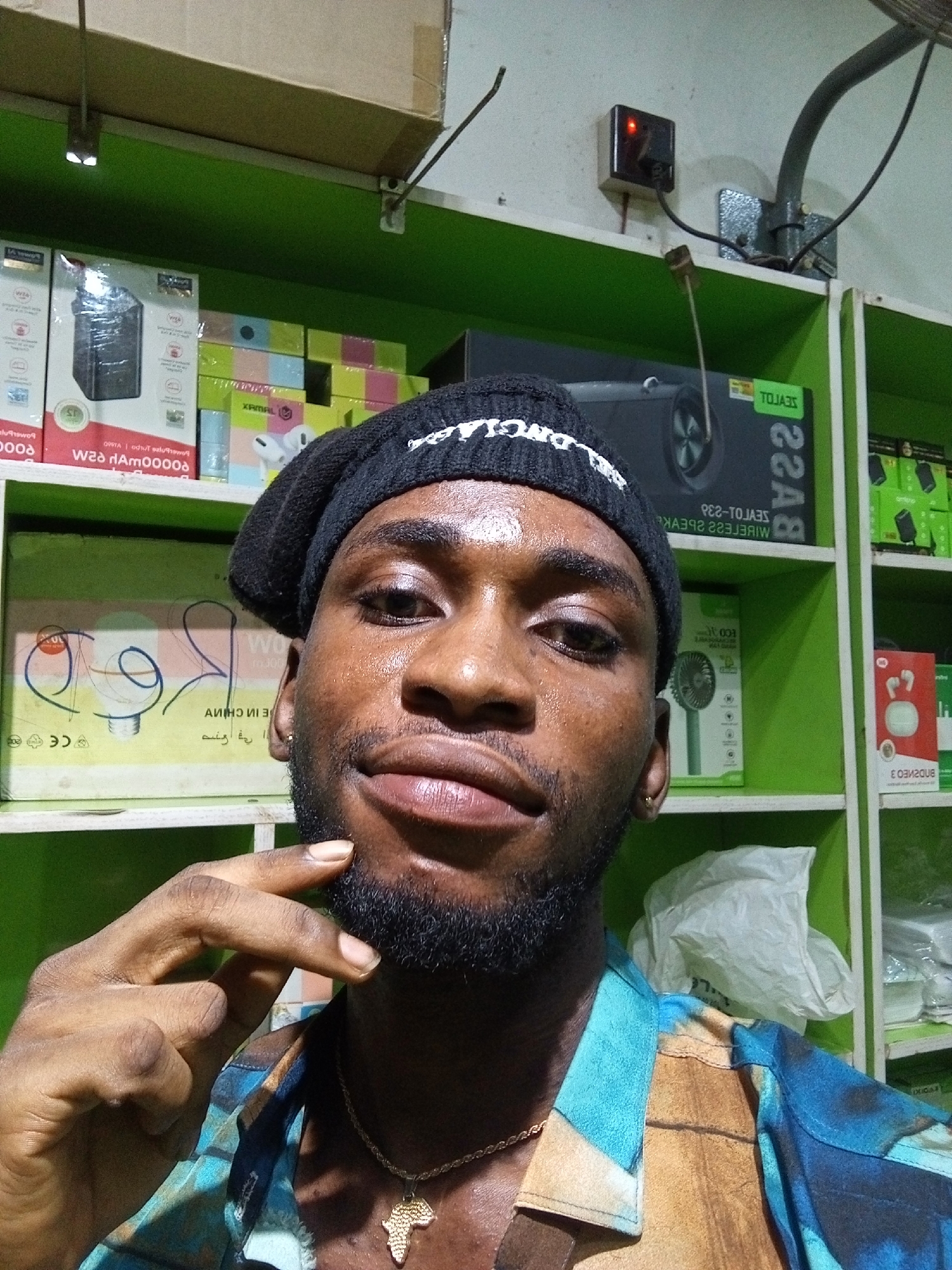 In a retail setting, a young Black man with a beard and a black beanie is captured in a close-up, chest-up selfie. He is wearing a vibrant, multi-colored shirt and a gold chain with an Africa-shaped pendant. His right hand is raised to his chin, and he is looking directly at the camera. Behind him, shelves are stocked with various electronic products in their packaging, including speakers and fans, suggesting a store in Lokoja, Nigeria, that sells electronics. The green shelves are typical of retail display units.