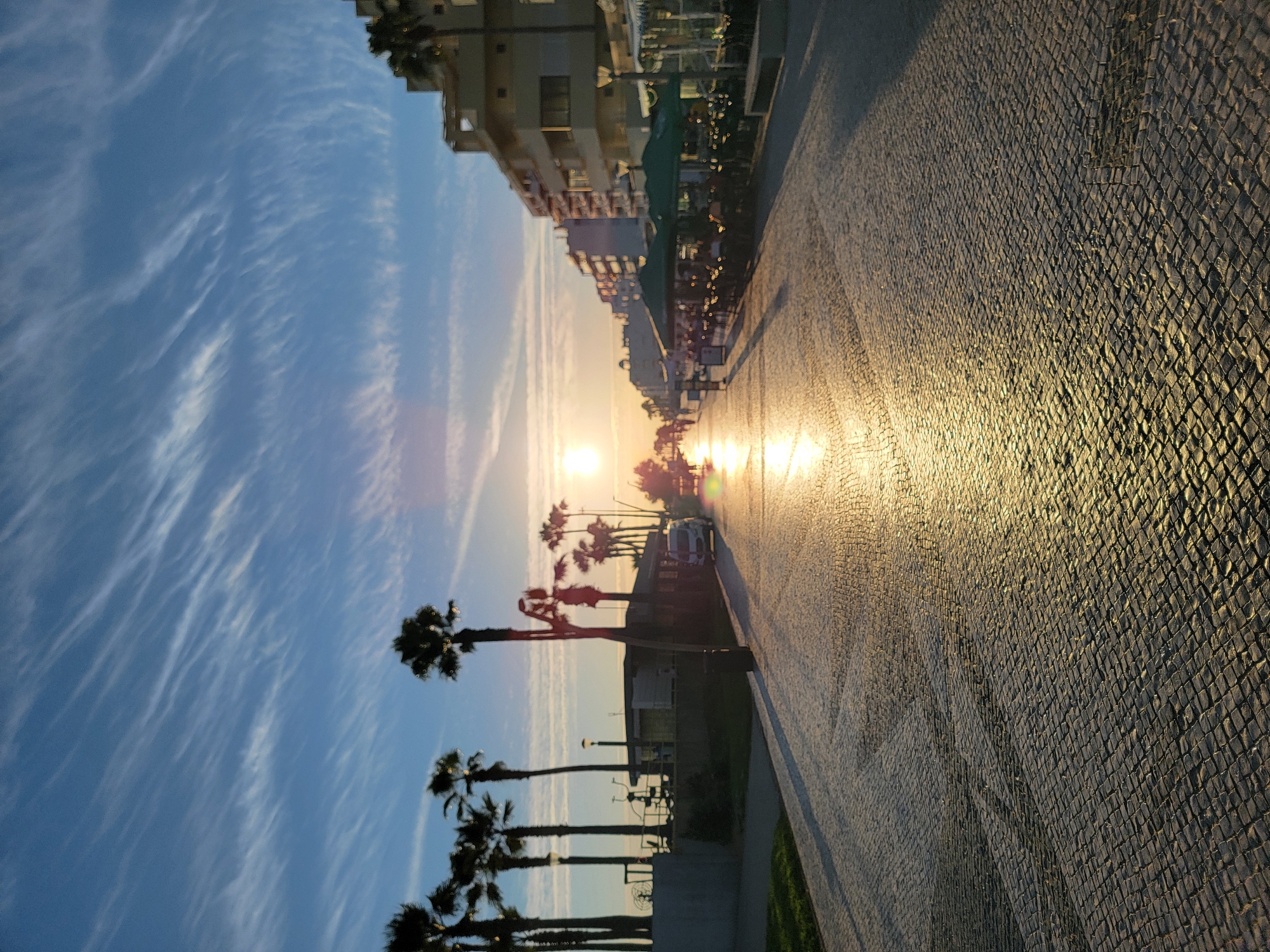 This is a wide-angle, eye-level, vertical, full-shot photograph of a paved promenade in Quarteira, Portugal, at sunset. The sun, low on the horizon, casts a strong reflection across the cobblestone pavement. Several palm trees are visible on the left side of the promenade, silhouetted against the bright sky. On the right side, buildings with balconies and restaurants with green umbrellas line the street. The sky is blue with wispy white clouds. A white van is parked on the right side of the promenade. Several people are visible walking in the distance.