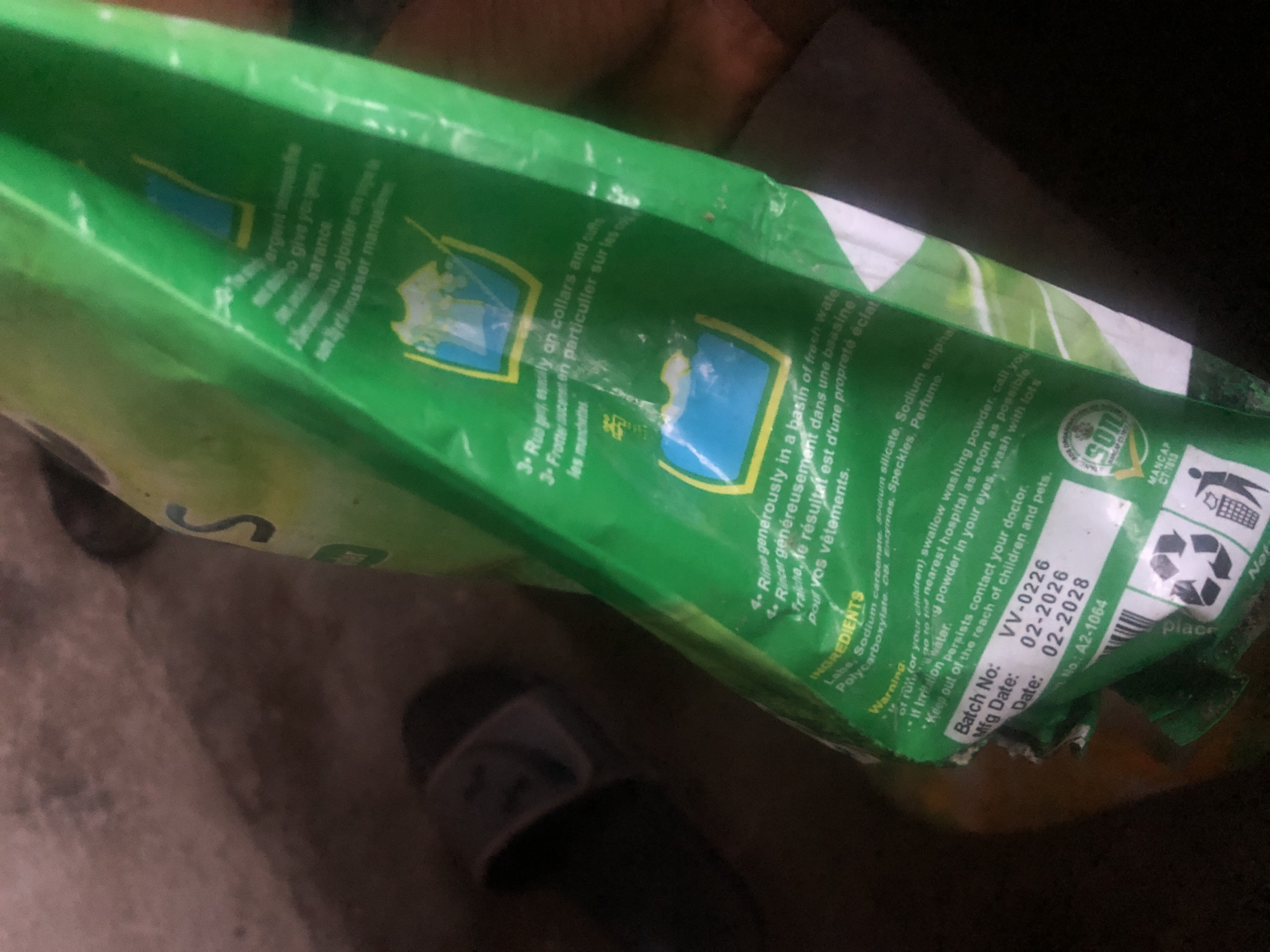 The image shows a close-up of a green plastic detergent bag. The bag is creased and partially unzipped. The text on the bag is in English and French and provides instructions for using the detergent, as well as a list of ingredients and a warning. The bag appears to be lying on a rough, textured surface, possibly a concrete floor. A shadow is cast across the surface, obscuring some details of the environment. The lighting suggests it is indoors or in a shaded outdoor area. The image is blurry and out of focus in some areas, but key details like text and product imagery are still discernible. The specific location in Jalingo, Nigeria, is not directly indicated by any visual cues in the image itself.