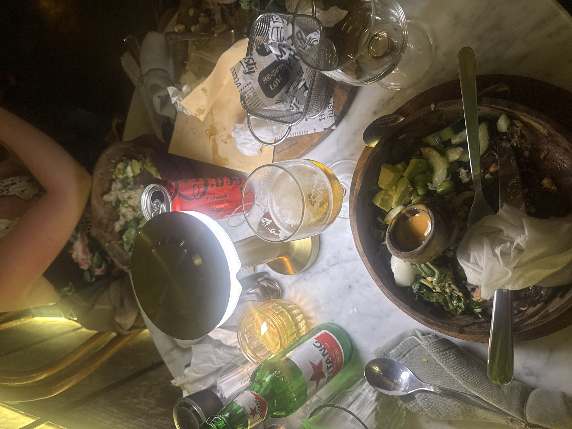 The image captures a lively table scene, likely in the evening, at a restaurant or bar in Ubud District, Indonesia. The setting features a marble-top table laden with various items, suggesting a recently concluded or ongoing meal with drinks.
**Subjects:** On the left, a person's arm and a floral-patterned top are visible. The table is crowded with objects including two wooden bowls, one containing remnants of a salad with avocado, cucumber, and edamame, and a small dressing cup. The other bowl is mostly empty. There are several glasses, including a beer glass containing some amber liquid and another empty one, along with various metal utensils (forks, spoons, knife) and crumpled napkins. Beverages include a green Bintang Pilsner beer bottle and a red Coca-Cola can. A modern table lamp provides bright illumination, complemented by the warm glow of a lit candle in a glass holder. A small metal basket lined with patterned paper sits on the table, alongside what appears to be a used paper bag.
**Activity or Event:** The scene depicts the aftermath of a casual dining experience, characterized by consumed food and drinks, indicating a relaxed social gathering.
**Notable Details:** The artificial lighting from the lamp and candle, combined with the general dimness, strongly suggests that the photo was taken at night or during the evening. The presence of a Bintang Pilsner bottle is a clear location cue, confirming the Indonesian context (Ubud District). The overall impression is one of casual enjoyment and a pleasant social atmosphere.
**Visible Text:** Text includes "BINTANG" and "Pilsner" on the green bottle, "FIFA WORLD CUP" and "FIFA" on the red Coca-Cola can. Paper items show "HAND-CRAFTED," "LOVE," "just," "VLLIC," "sty," "B," and "104." "HPICK" is also visible, possibly on a dispenser.
