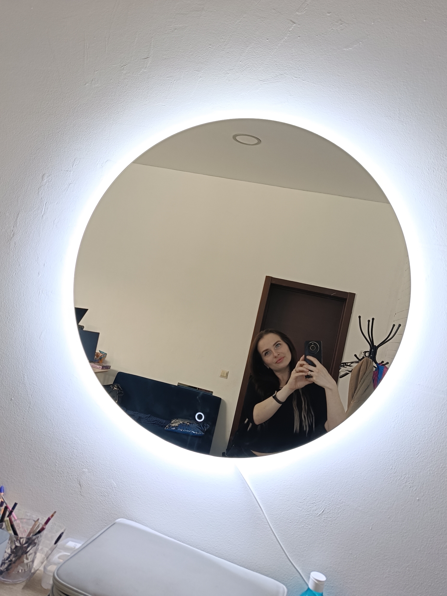 A young woman is taking a selfie in a round, illuminated mirror. The reflection reveals elements of a room, including a dark-colored door, a coat rack with clothing, and part of a couch in the background. Below the mirror, makeup brushes and a case are visible. The location is Chaikovsky, Russia.