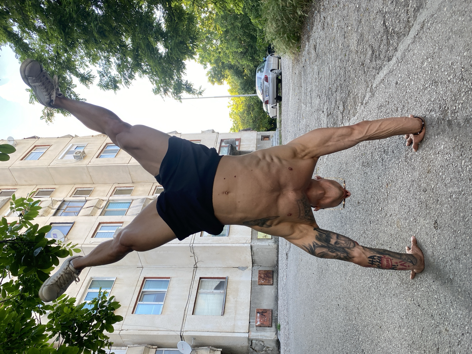 A muscular male individual performs a handstand on a paved surface in Pleven, Bulgaria. He is wearing black shorts and white athletic shoes, with his legs extended upwards and slightly apart. His torso is exposed, revealing a navel piercing and several moles/birthmarks. Distinctive tattoos are visible on both forearms; the left forearm features a portrait resembling Tony Montana and text including "HA HA INSANE," while the right forearm displays intricate dark designs. A dark beaded bracelet is on his right wrist, and glasses are visible hanging around the back of his neck.
The setting is an urban outdoor environment. To the viewer's left, a multi-story, light-colored apartment building with numerous windows and several satellite dishes is visible. The building exhibits signs of age with some wear on its facade and rusting metal panels at its base. Green tree foliage partially obscures the upper left portion of the building. To the viewer's right, a dense area of green trees and vegetation flanks a dirt or gravel path. A light-colored sedan, identified as a Renault Laguna II with Bulgarian license plate "T 6103 AK" (indicating registration in Pleven), is parked facing away from the viewer, with another white car partially visible behind it. A light pole stands between the parked cars and the trees. The ground beneath the individual is a rough asphalt or concrete surface with scattered small stones and a faint white line on the right. The sky is bright with some cloud cover.
