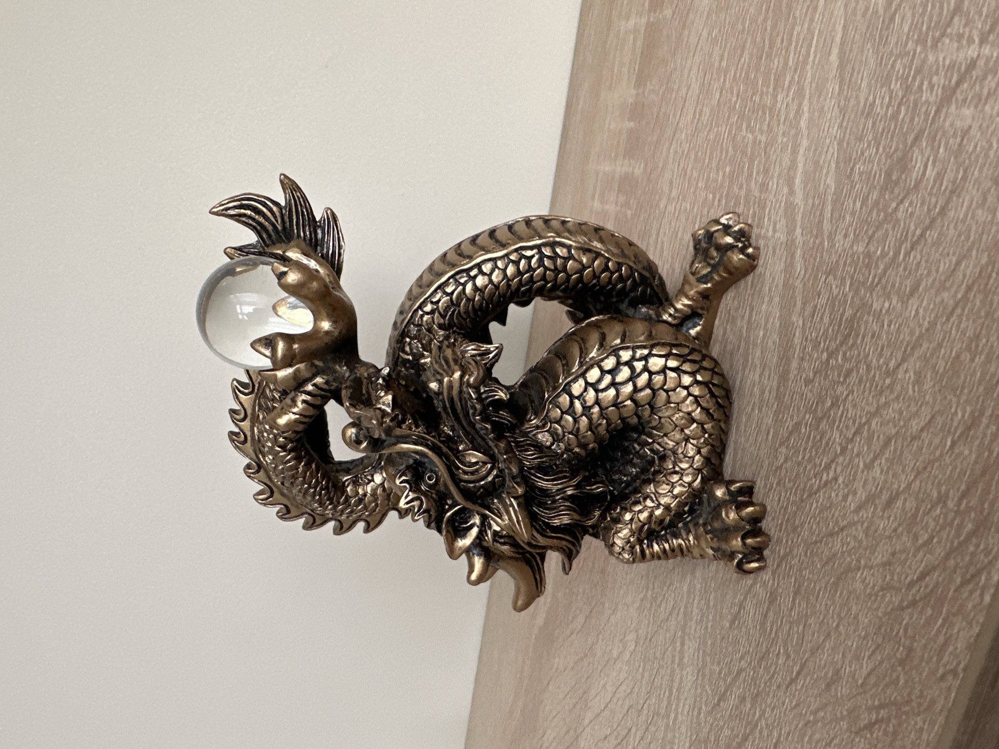 A close-up shot presents a detailed metallic dragon figurine. The dragon, finished in a brassy or golden hue, is depicted in a coiled pose, clutching a clear, spherical glass ball with its front claws. The figure exhibits intricate detailing, with scales, claws, teeth, and a spiky mane. The background of the image is split between a light cream or white wall and a section of wood paneling with a light, grainy finish. The positioning of the dragon suggests it is situated on the corner where the wall and paneling meet. No details indicate the location being Oryol, Russia.