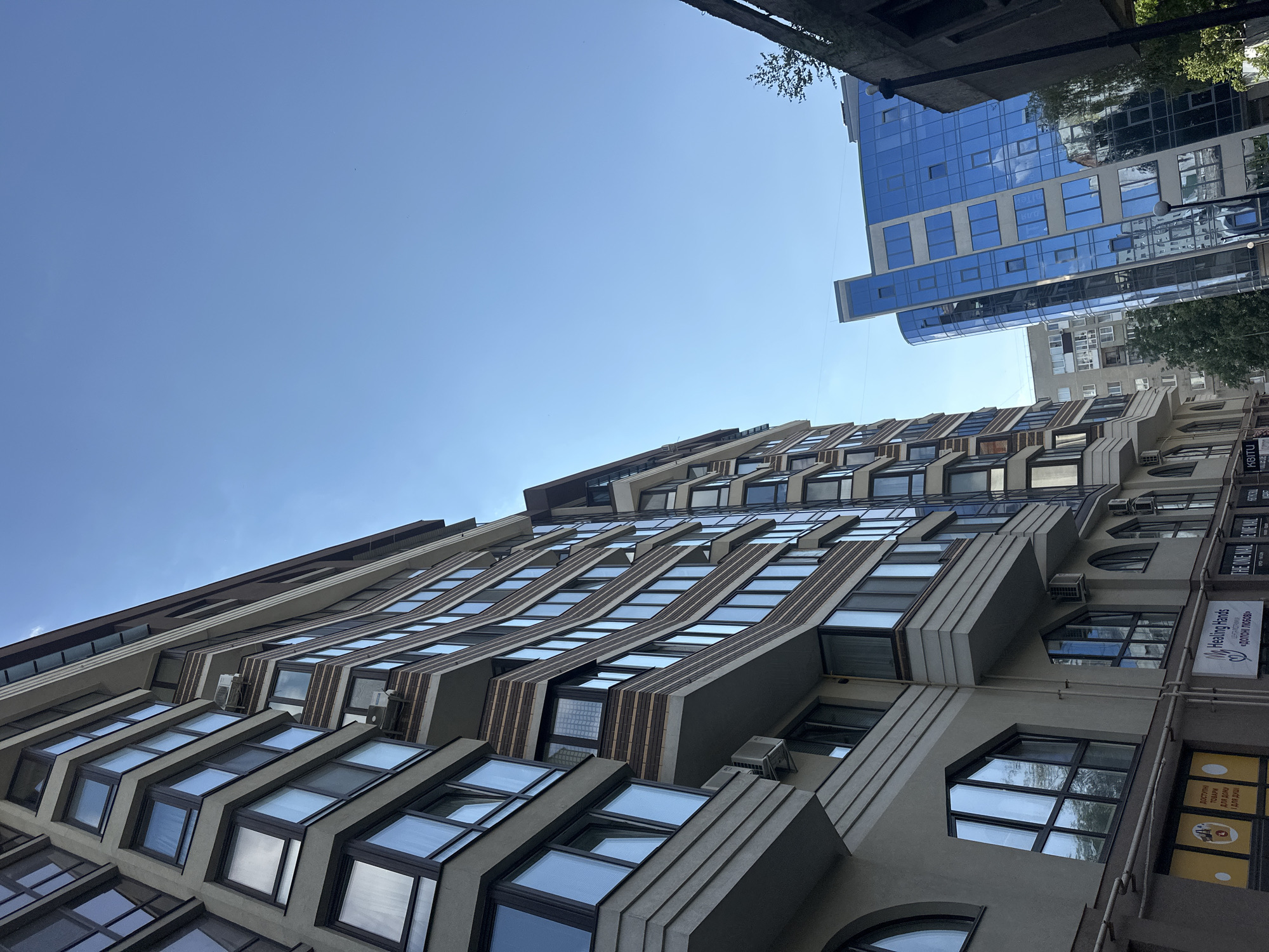 The image captures a low-angle view of a street in Ternopil, Ukraine, dominated by modern architecture. The primary focus is on two multi-story buildings. On the left, a beige-colored apartment building with numerous angular windows and balconies stretches upwards, its facade featuring decorative brickwork. Air conditioning units are visible on some of the balconies. To the right, a striking blue glass-fronted building curves outwards, reflecting the clear blue sky and surrounding structures. In the foreground, at the base of the beige building, shop signs are partially visible, including one that reads "Healing Hands." The environment appears to be a bright, sunny day with a clear sky.
