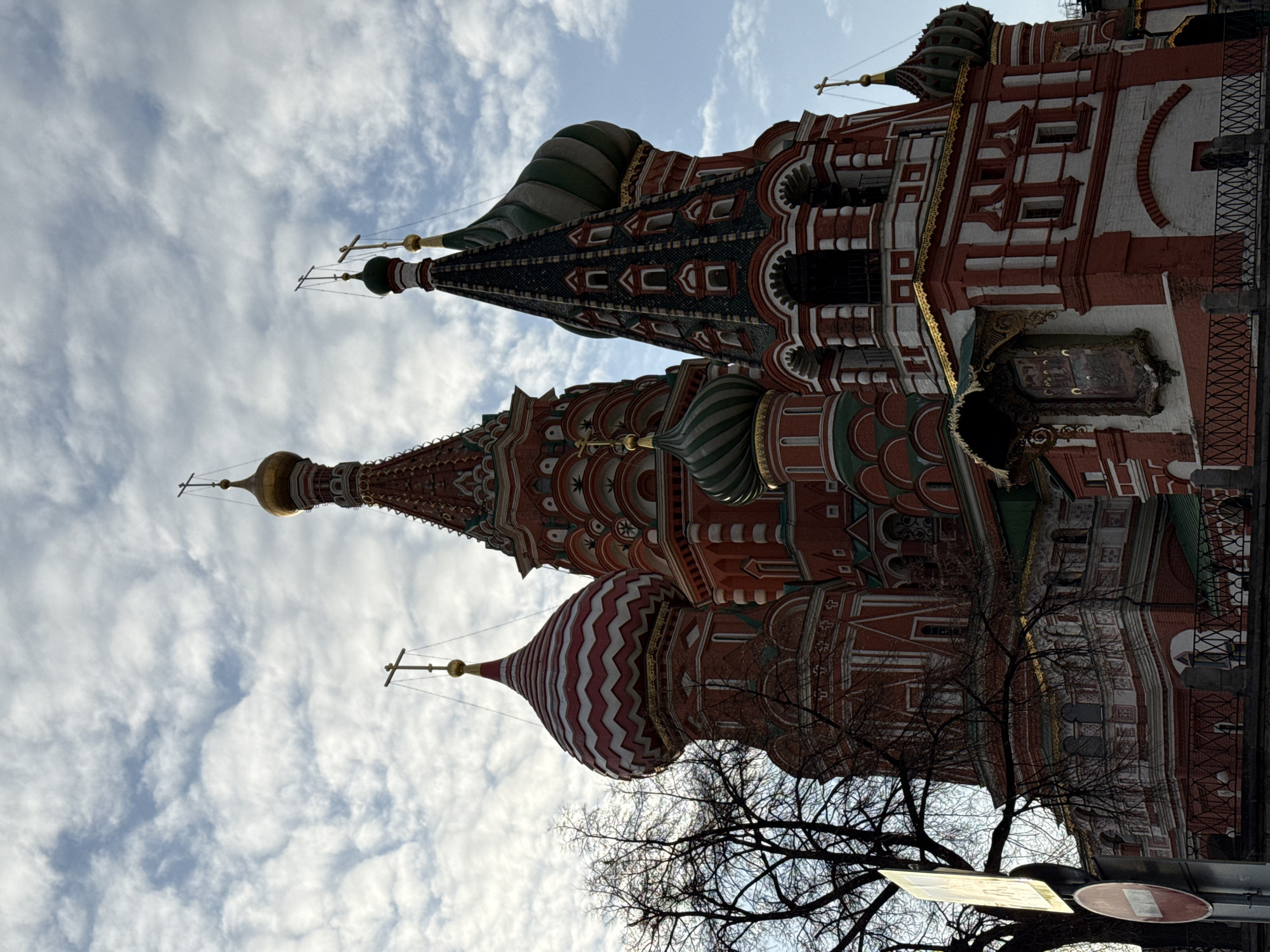 The image depicts a detailed, upward-angled view of St. Basil's Cathedral in Red Square, Moscow, Russia, under a partially cloudy sky. The cathedral, a prominent architectural subject, features multiple ornate domes and tent-like roofs.
The structure is predominantly red brick with intricate white, green, and gold decorative elements. Visible elements include:
* A tall, central tent-like spire topped with a golden onion dome and an Orthodox cross.
* To the left, a large onion dome with a distinctive red and white zigzag pattern, also crowned with an Orthodox cross.
* Various other domes and roofs, including a dark green conical roof with multiple small white-framed windows, and smaller green and white striped onion domes. All visible domes are topped with golden Orthodox crosses.
* An outdoor icon or religious painting, framed and positioned under a small decorative canopy, is affixed to a lower section of the cathedral wall.
* Gold detailing highlights cornices and architectural contours across the building's facade.
In the lower left foreground, a bare deciduous tree with dark, fine branches partially obstructs the view of the cathedral's base. Below and to the left of the tree, a street sign is visible, featuring a circular red 'No Entry' symbol (a white horizontal bar on a red background). A dark metal fence or railing runs horizontally across the lower part of the frame, separating the foreground from the cathedral's base. The sky above is filled with scattered white and gray stratocumulus clouds, allowing glimpses of lighter blue sky. No individuals are visible within the frame.