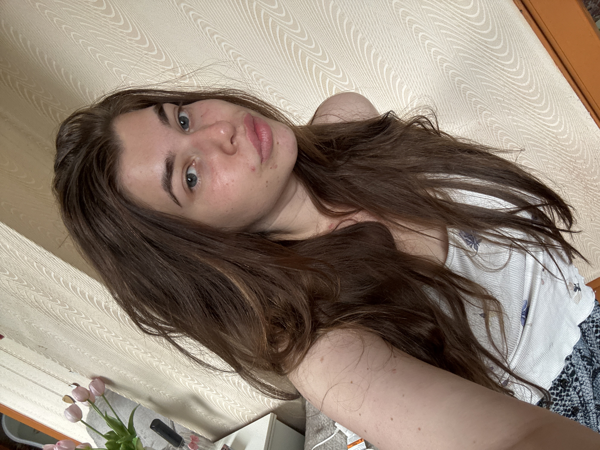 A young woman with long, dark brown hair and blue eyes is shown from the chest up, looking towards the camera. She is wearing a white tank top with a floral pattern and a dark, patterned bottom. The background consists of a textured, cream-colored wall with a wave-like pattern, and a sliver of a wooden surface is visible at the bottom right. To the left, a bedside table with a vase of pink tulips and a black object, possibly a phone or camera, can be seen. The woman has freckles on her cheeks and appears to have some redness around her nose and cheeks. The lighting suggests it is daytime, possibly with natural light coming from the side. The scene appears to be an indoor setting, likely a bedroom. The city of Мещёры, Russia, is not visually identifiable within the image.