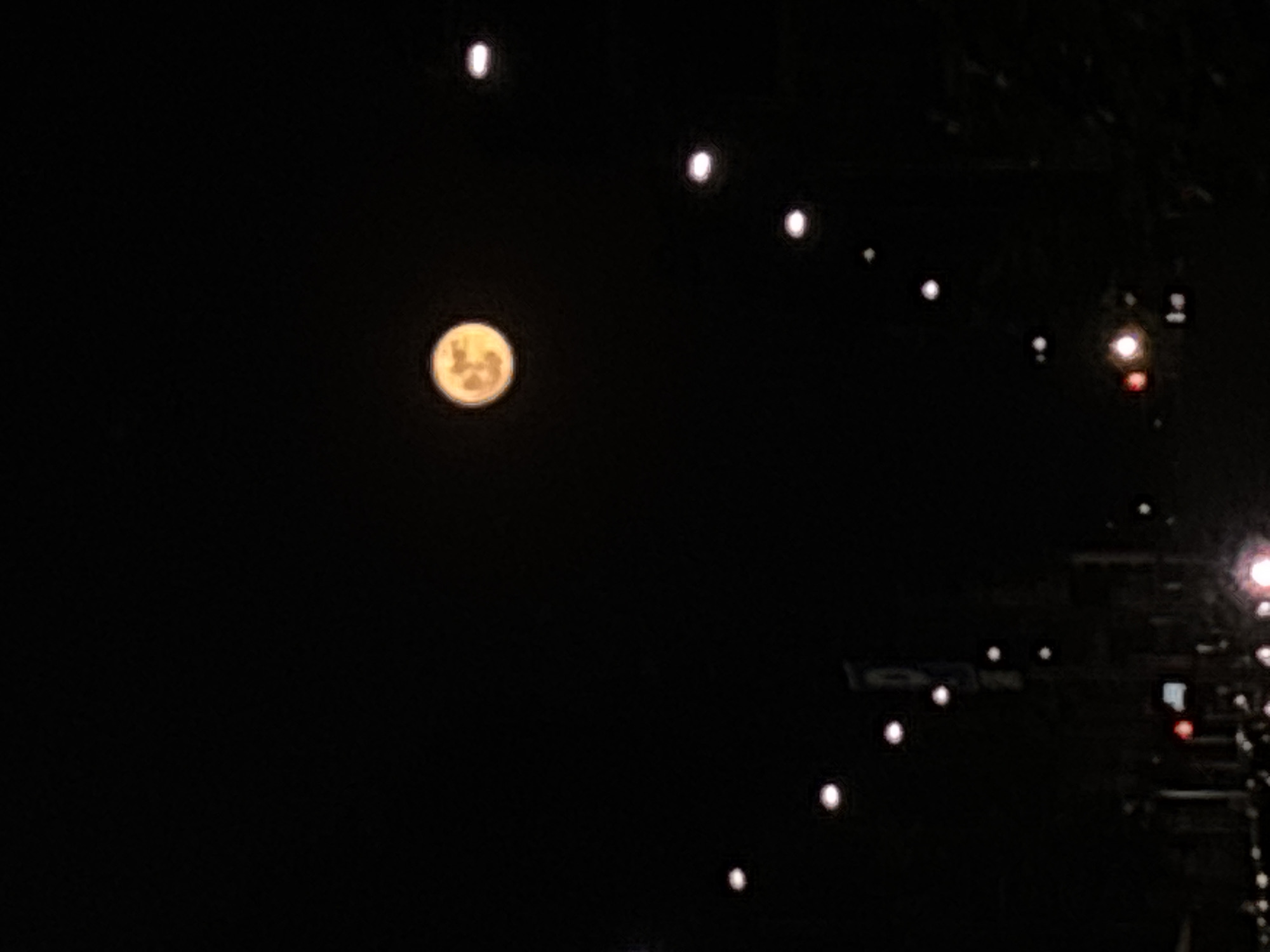 The image captures a night scene in Milan, Italy, dominated by a dark sky. The moon is a prominent feature, appearing round with visible surface details. A series of evenly spaced streetlights stretches across the upper and right side of the frame, indicating a street or bridge. A hint of building structures or other city elements can be seen blurred in the lower right corner.