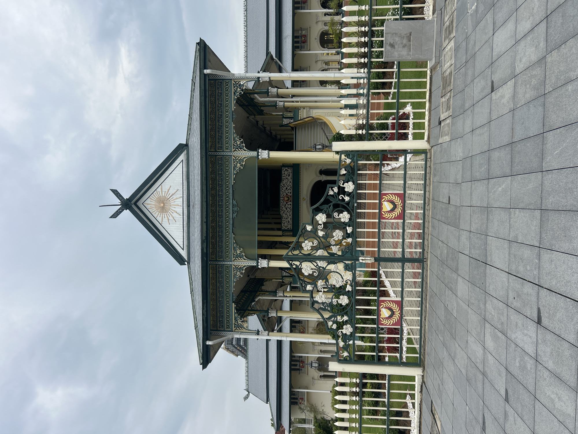 The image shows the main entrance to a grand, colonial-style building, likely a palace or government building, in Alor Setar, Malaysia. The building features a multi-tiered roof with dark grey tiles, a wide veranda supported by numerous columns painted yellow and white, and ornate green and gold decorative elements along the eaves and railing. A prominent triangular gable at the front of the roof displays a sunburst motif within a decorative frame and a crest at the apex.
In front of the building is a decorative green metal gate with intricate scrollwork featuring floral motifs. The gate is flanked by a white picket fence, with the gate and fence section at the entrance to the building having a more substantial and decorative appearance. Behind the fence, a neatly manicured lawn is visible, interspersed with some small red and white flowers and white gravel.
The foreground consists of a grey paved walkway made of large rectangular tiles, with visible seams. The sky is overcast and cloudy, suggesting a dull or perhaps rainy day. No people are visible in the image. The overall impression is one of formal architecture and well-maintained grounds. This building is likely the Istana Anak Bukit, the royal palace of the Sultan of Kedah, located in Alor Setar.